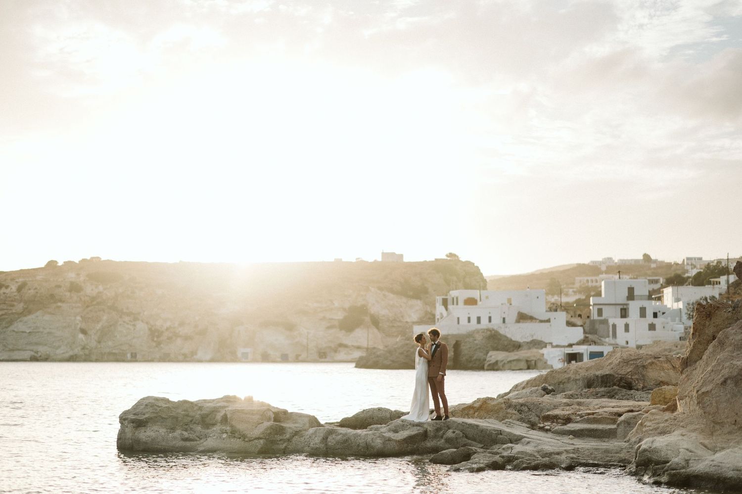 A silhouette stands on rocky coastal cliffs at sunset with white Mediterranean buildings in the background.