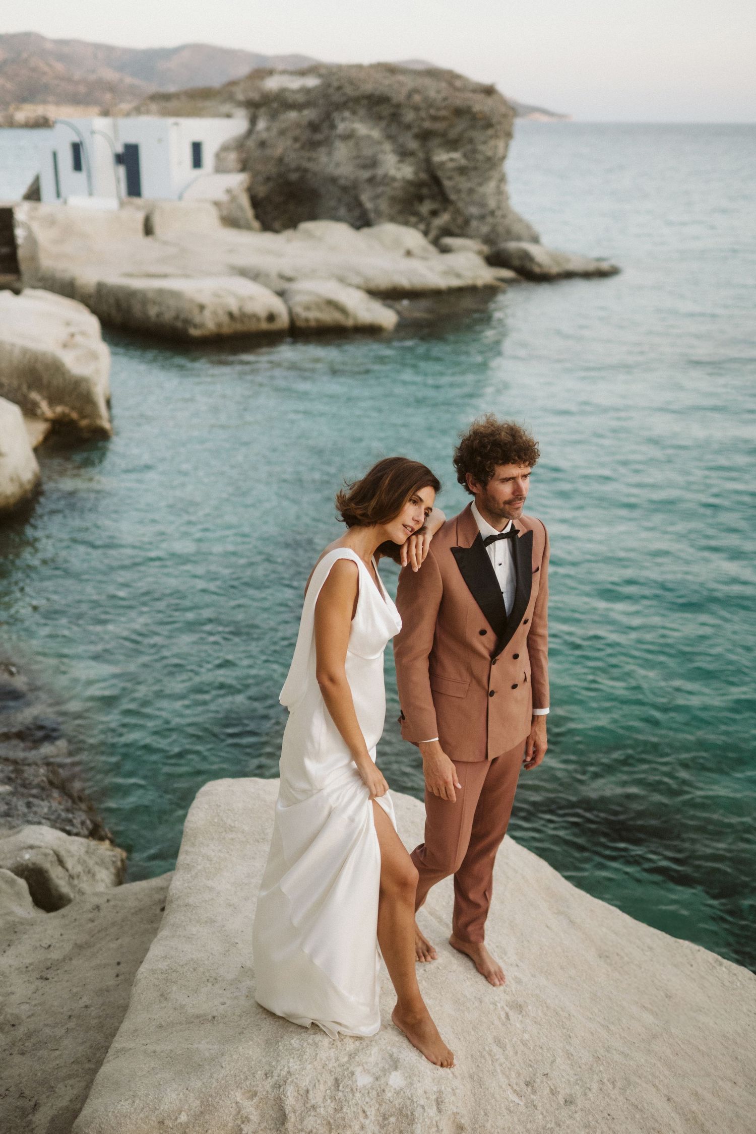 A couple poses barefoot on white coastal rocks near turquoise Mediterranean waters and traditional white buildings.