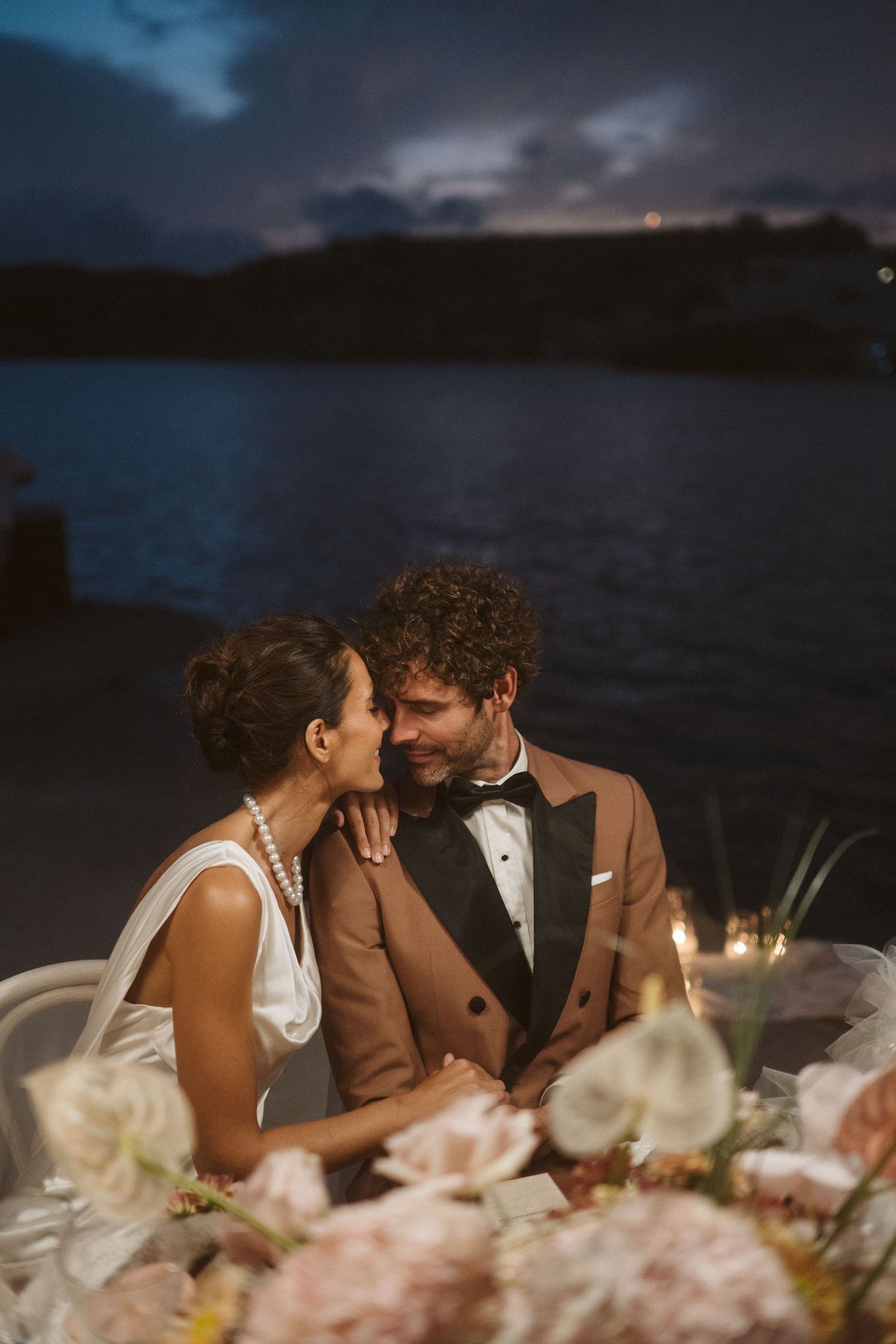 A romantic candlelit dinner setup by the water at dusk with floral decorations in soft pink tones.