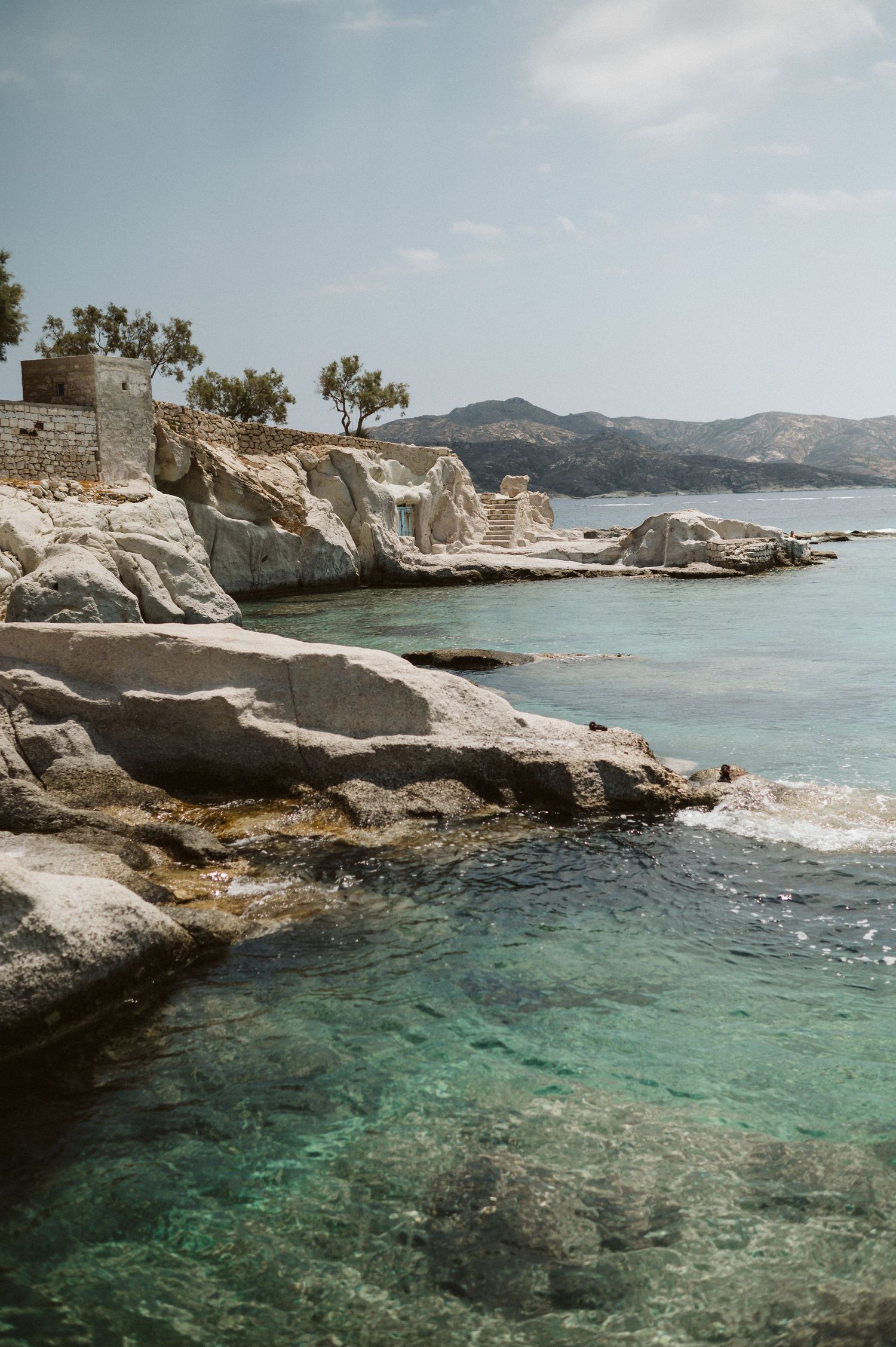 Rocky coastal landscape with turquoise Mediterranean waters and white limestone cliffs against mountainous backdrop.