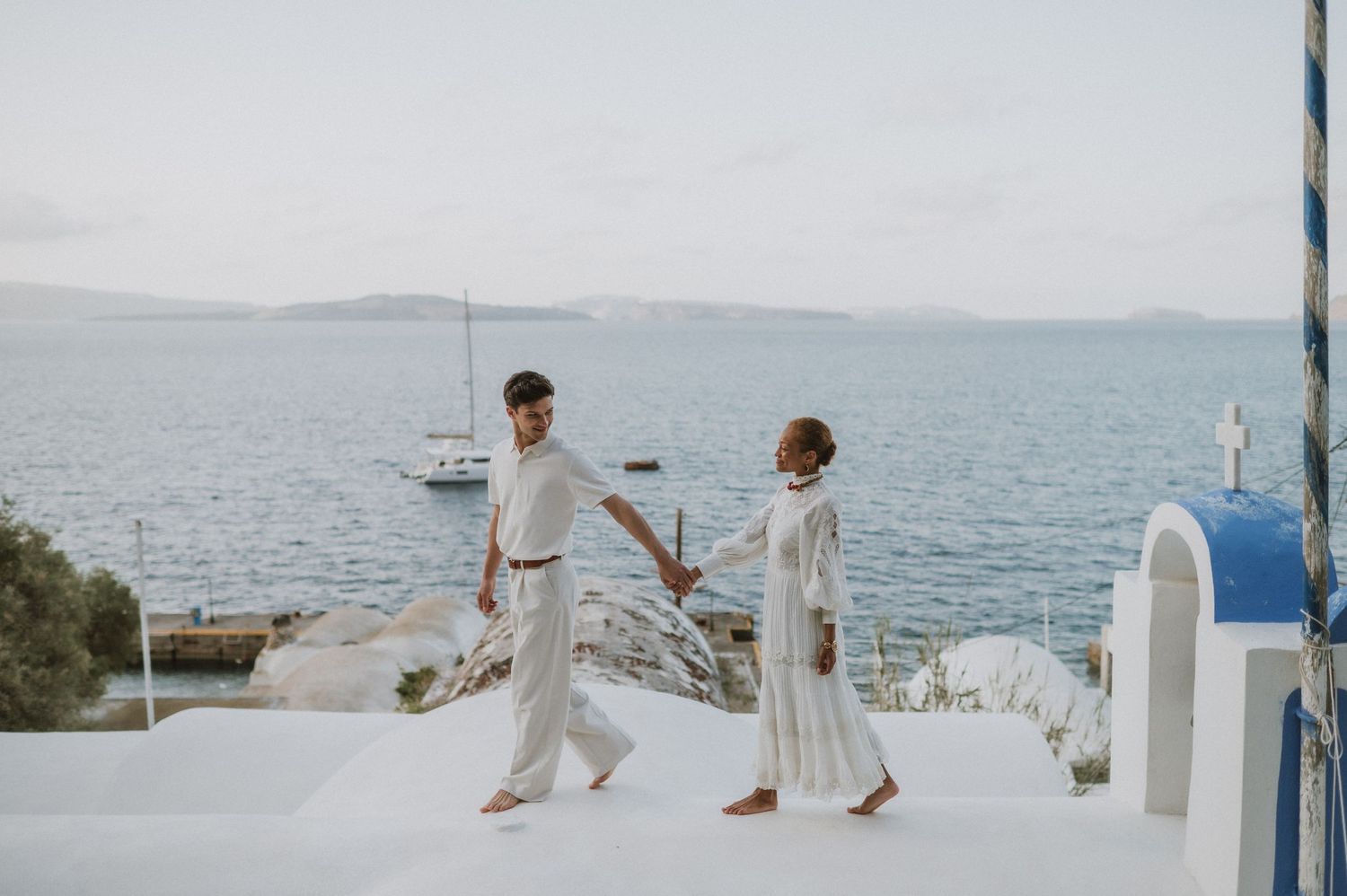 Two people in white outfits hold hands while walking on a Mediterranean terrace.