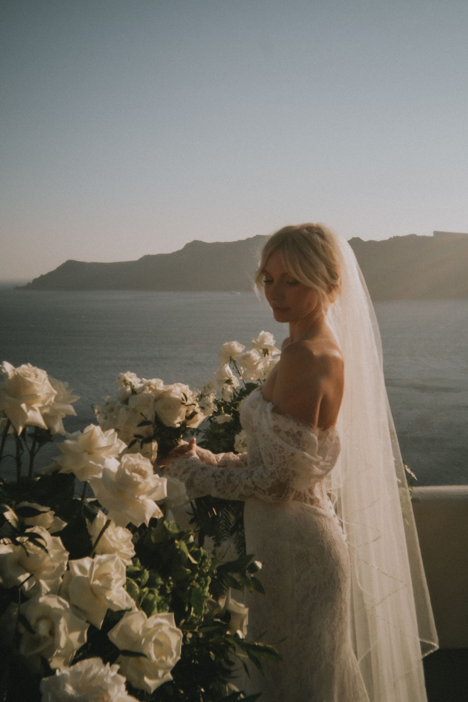 A bride in a white lace wedding dress stands against a scenic coastal mountain backdrop at sunset.