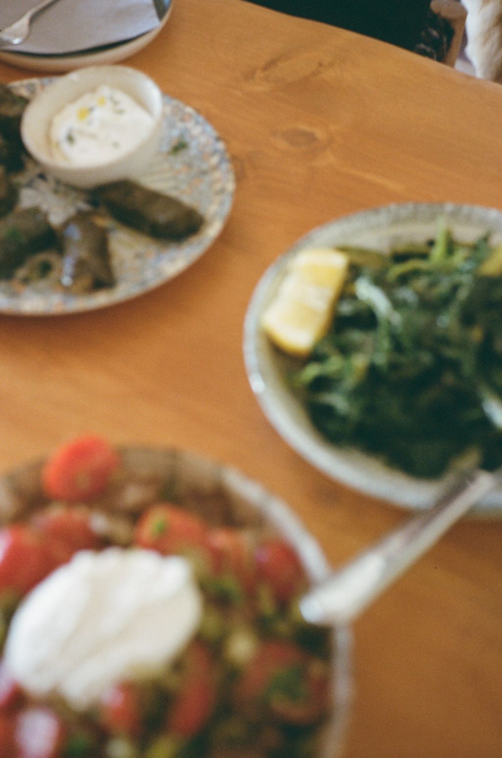 A blurred close-up view of plates with various dishes on a wooden table shows partial glimpses of food and tableware.