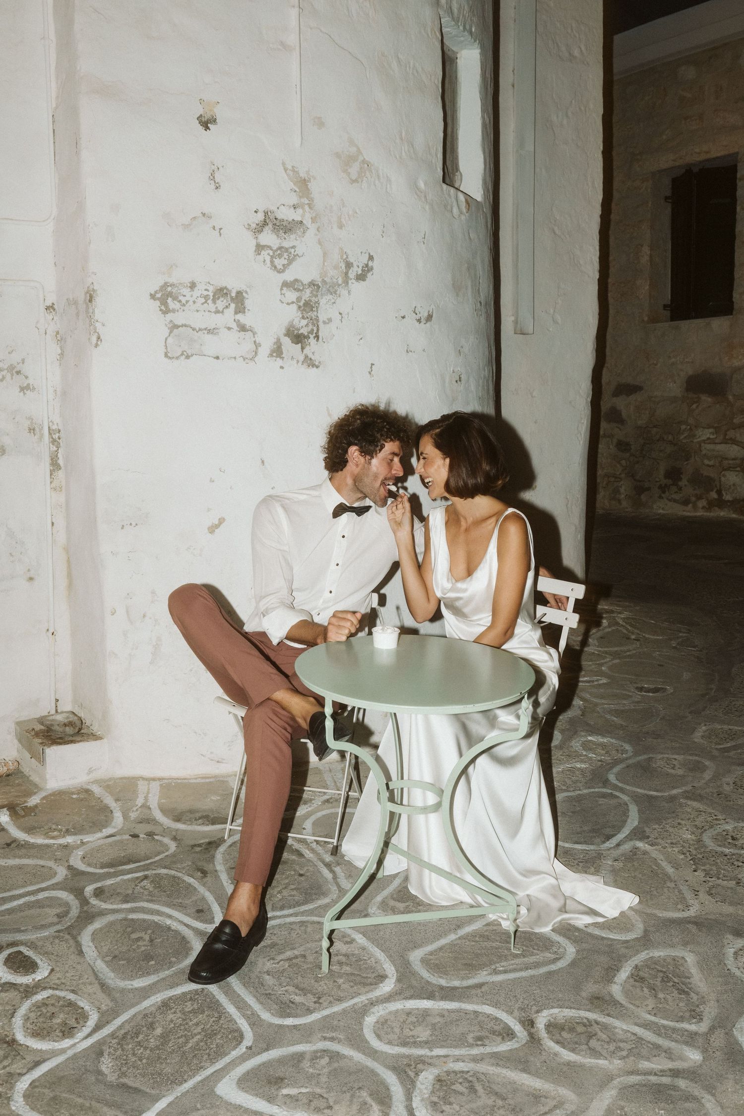 A romantic moment shared at a small bistro table with a weathered white wall and patterned stone floor background.