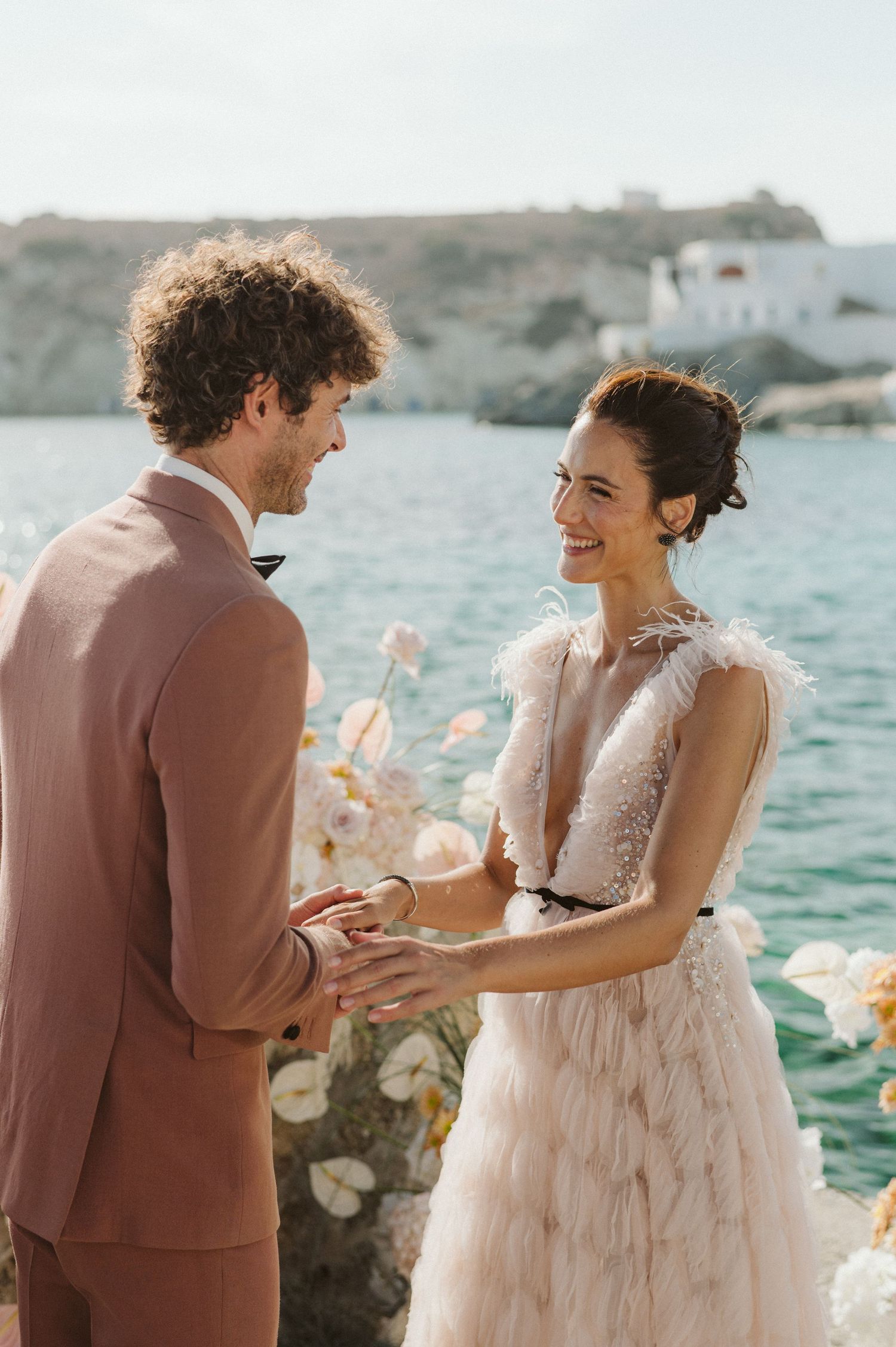 A couple in wedding attire share a romantic moment by the seaside, with blue water and white buildings in the background.