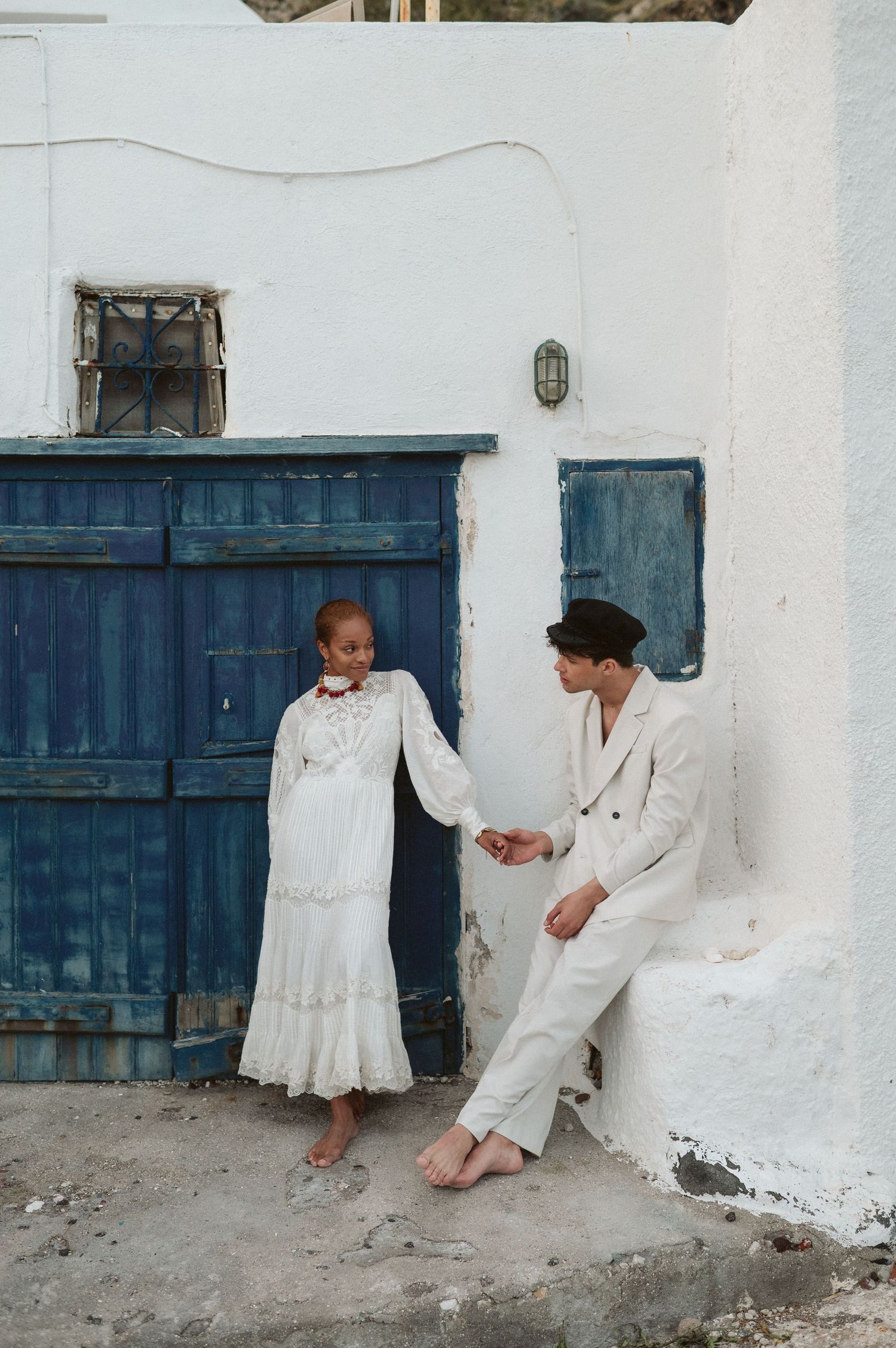 Two people in white outfits pose against a rustic whitewashed wall with blue doors.