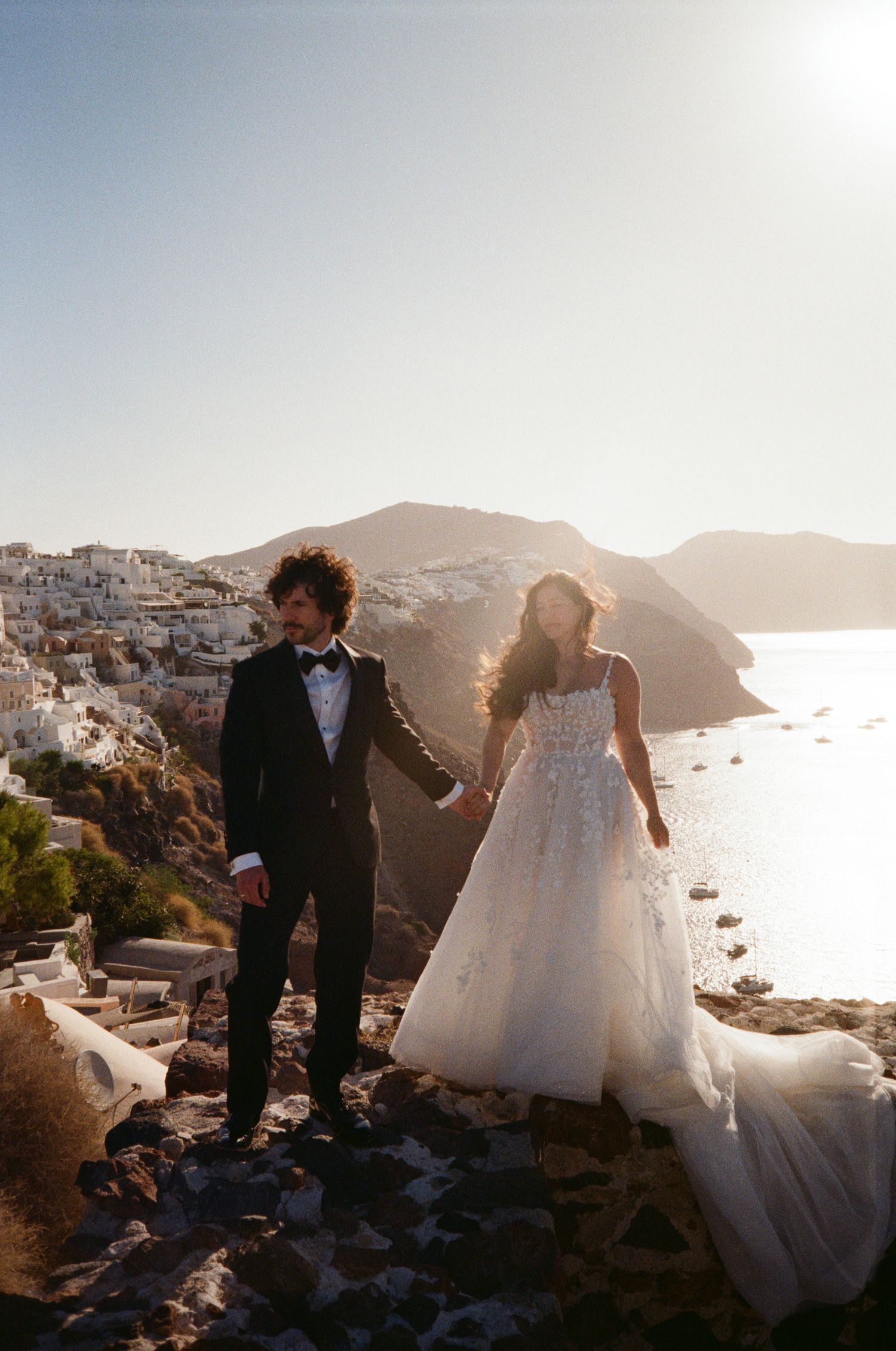 A couple in wedding attire stands on a cliff overlooking the white buildings and sea in Santorini, Greece at sunset.
