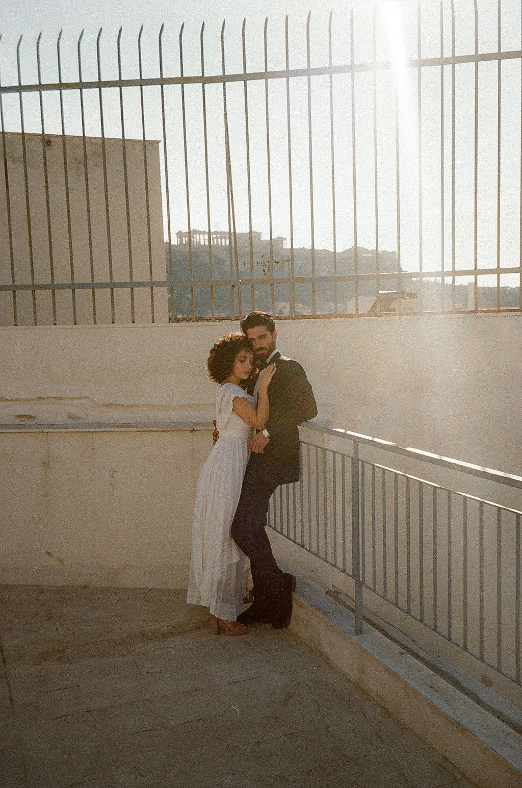 A romantic couple embraces against a railing in a sunlit industrial space with a dreamy, vintage aesthetic.