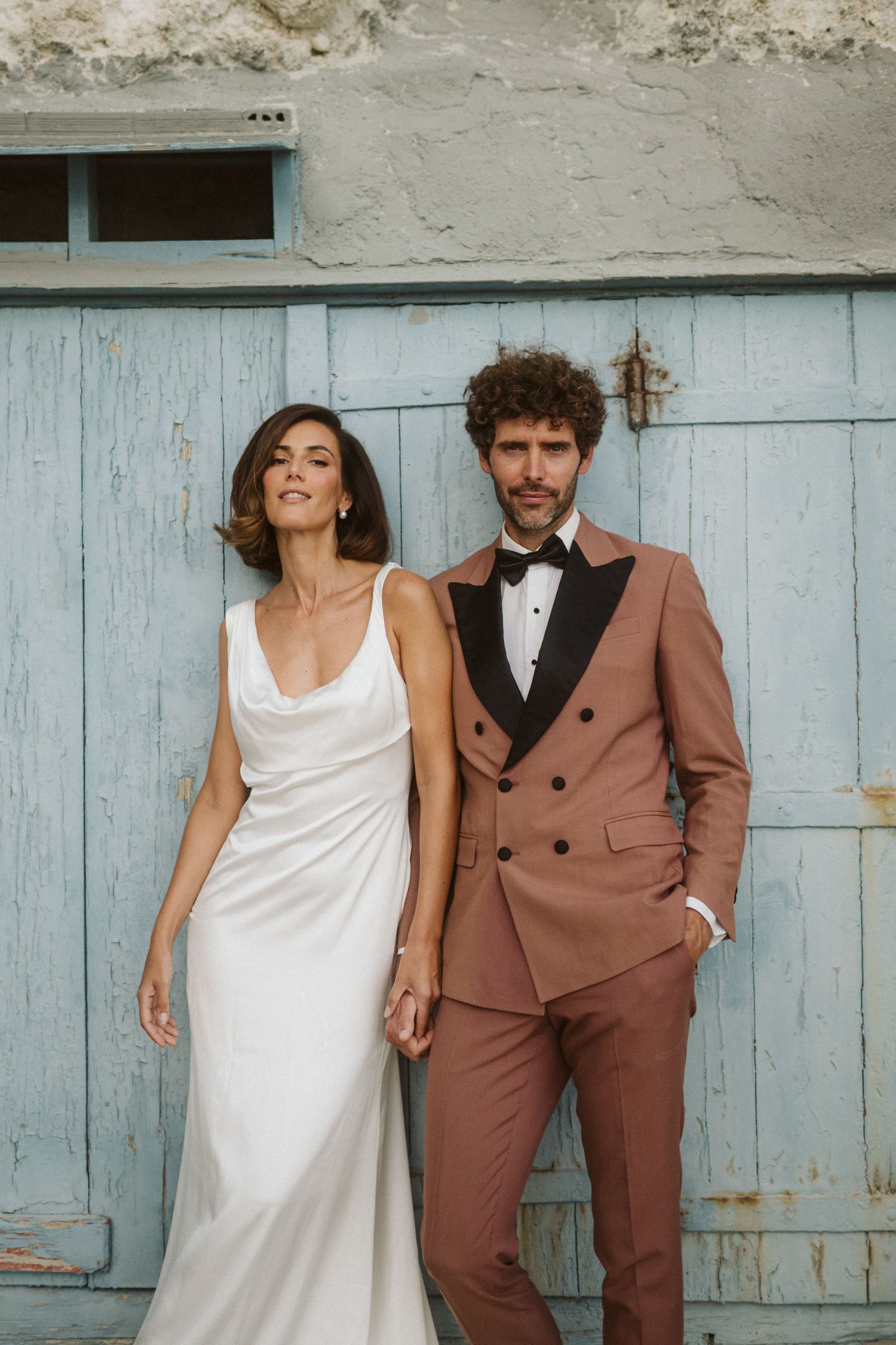 A stylish couple poses in wedding attire - white silk dress and dusty rose double-breasted suit - against a rustic blue door.