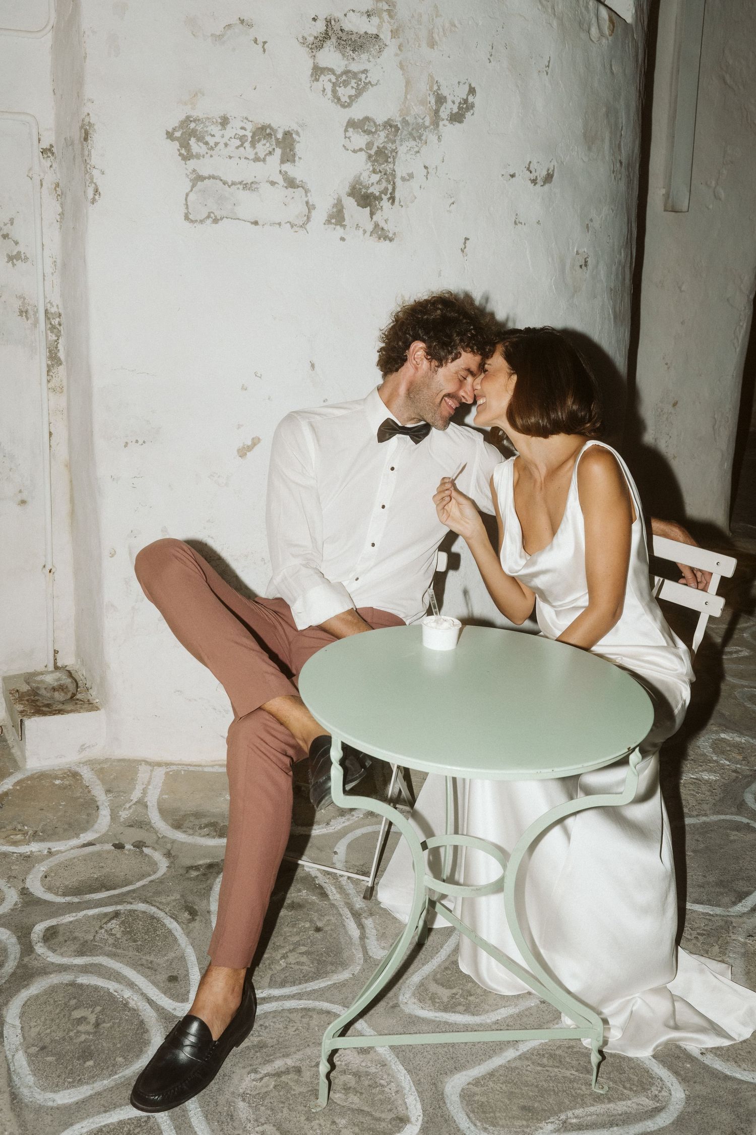 Romantic couple in white formal attire share intimate moment at small cafe table with textured floor pattern.