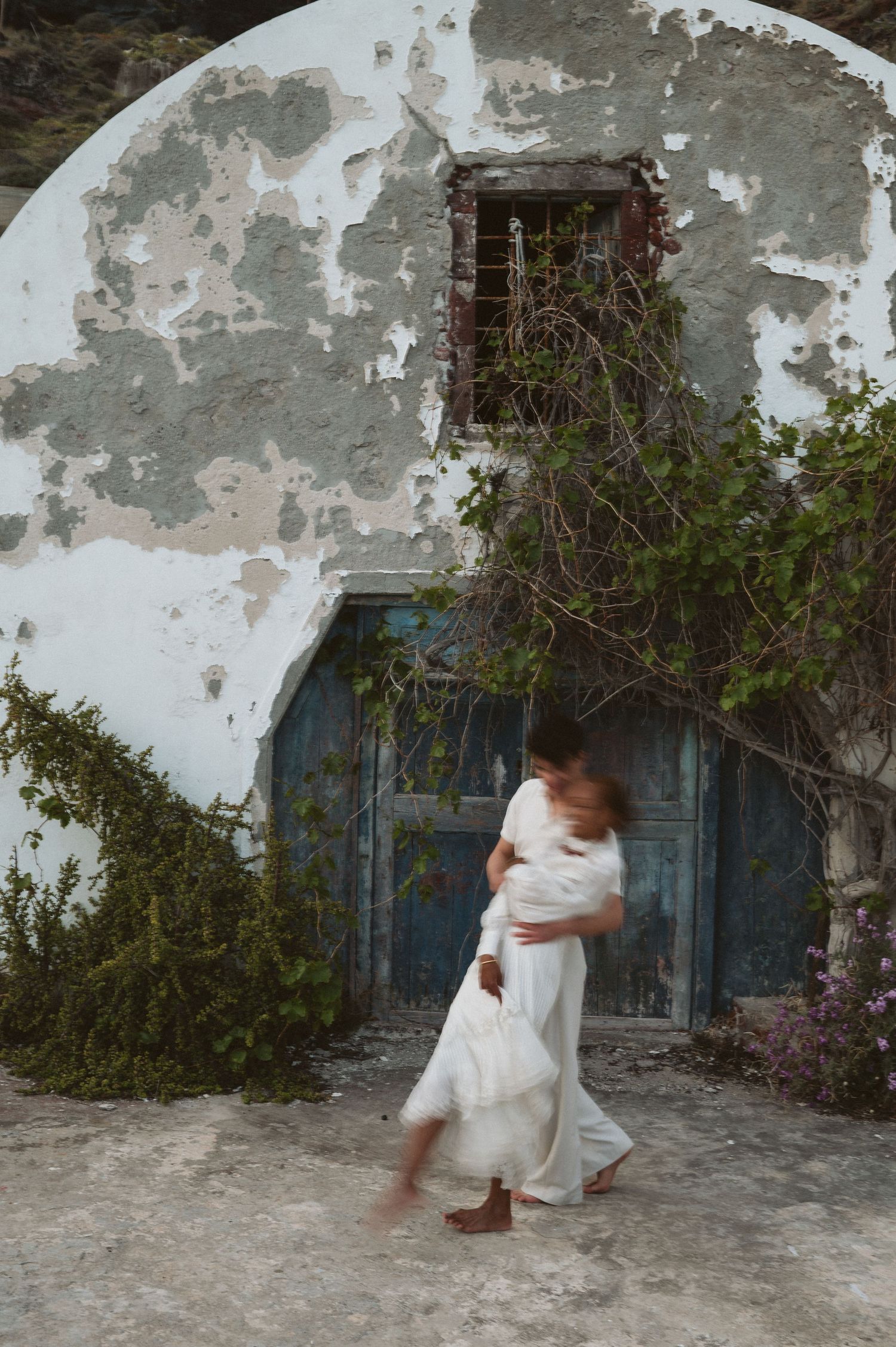 Figure in flowing white clothing dances in front of weathered blue door and white wall.