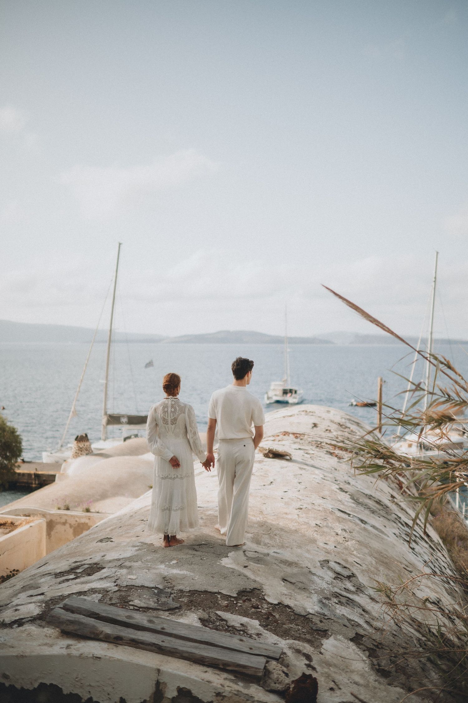 A couple in white clothing stands on a rocky coastal overlook with boats visible in the distance.