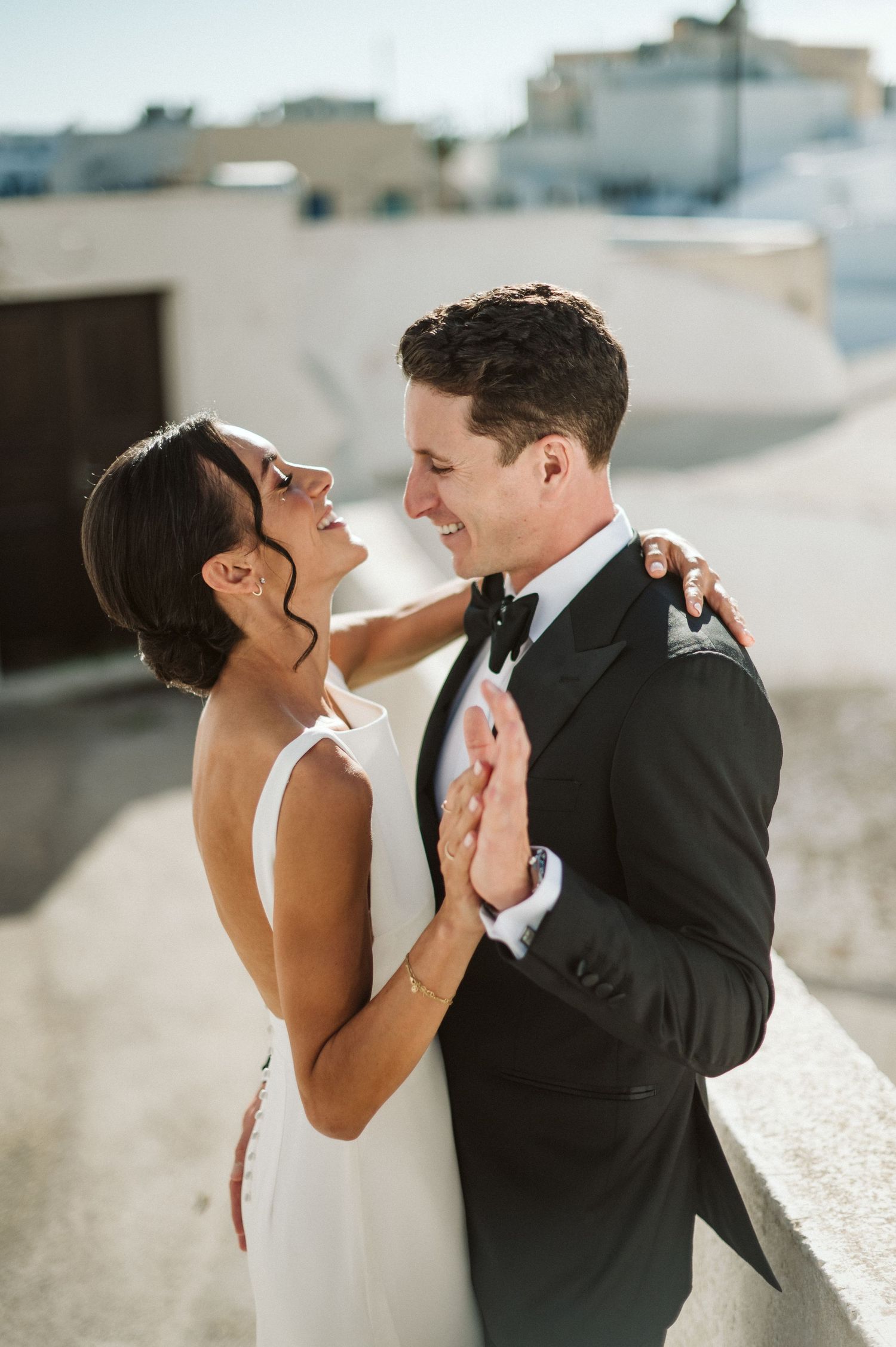 Romantic wedding portrait of a couple in formal attire embracing against a white building in Mediterranean sunlight.