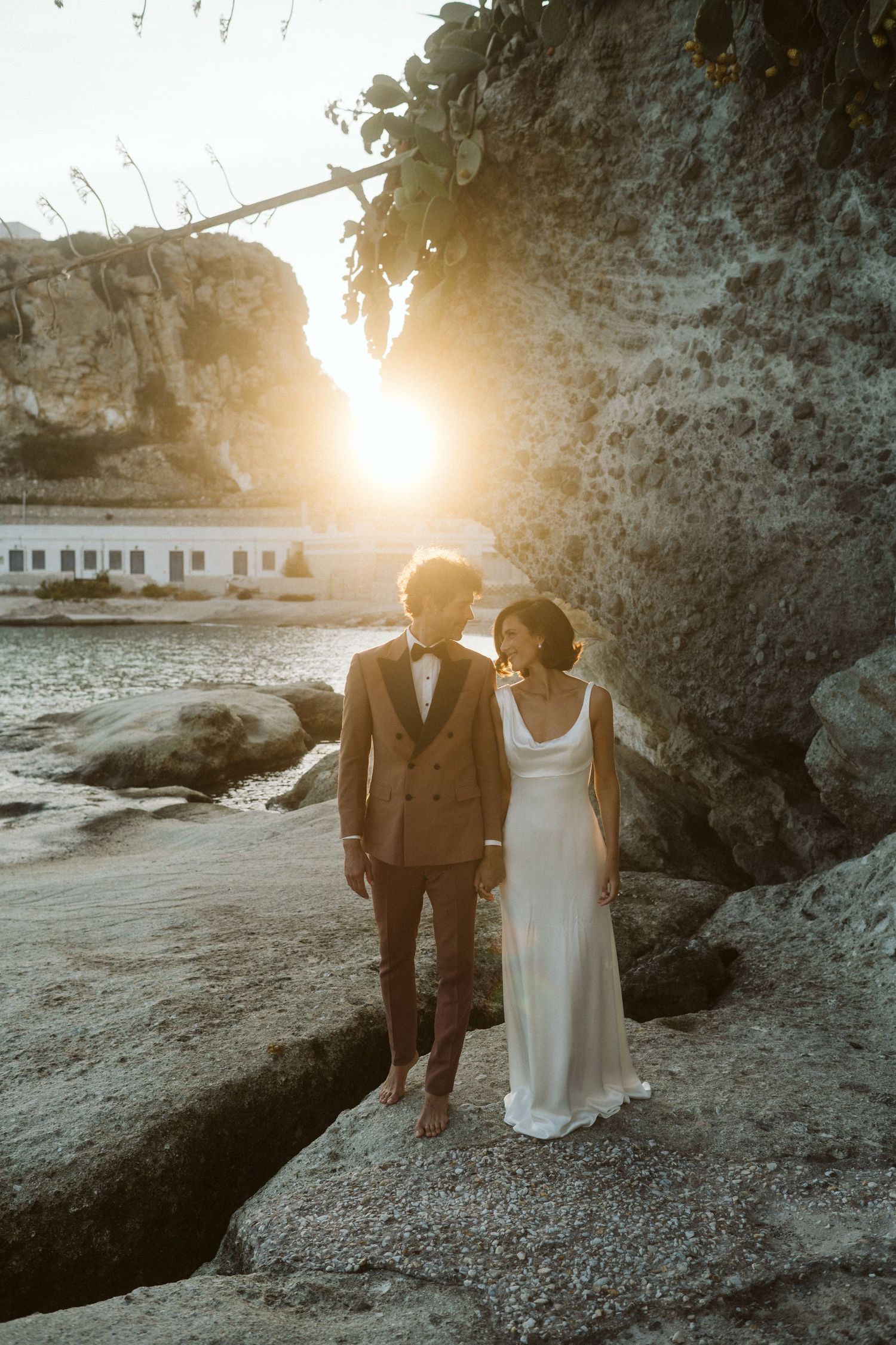 A couple in wedding attire stands together on coastal rocks during golden hour sunset near a cliff face.