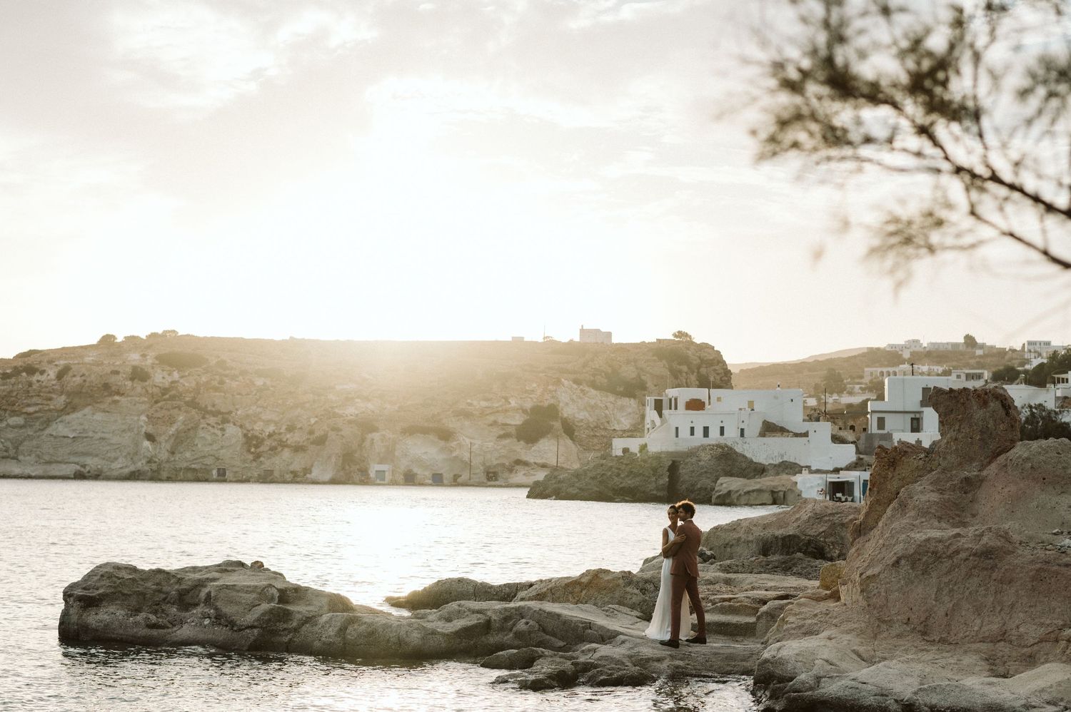 A silhouetted figure stands on rocky coastal outcrop at sunset with white Mediterranean buildings visible in background.