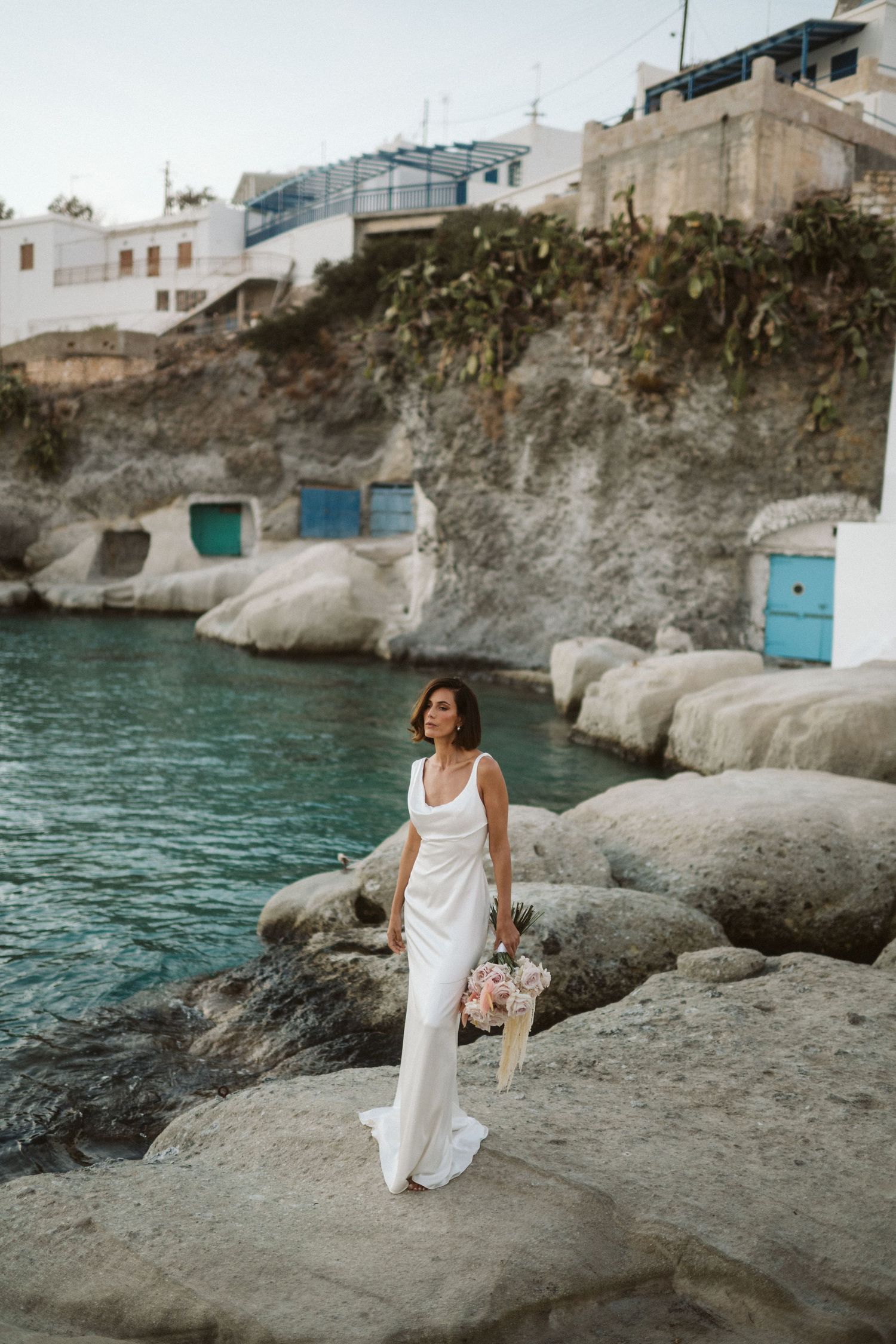 Woman in white dress stands on coastal rocks near turquoise water and white buildings in Mediterranean setting.