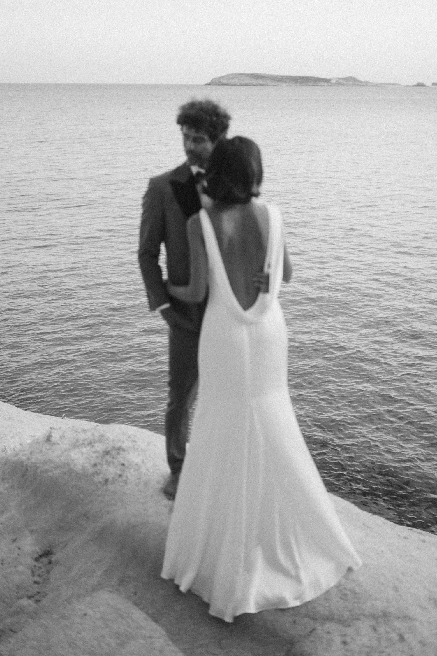 A wedding couple stands on a cliff edge overlooking a calm ocean and distant island in black and white photography.