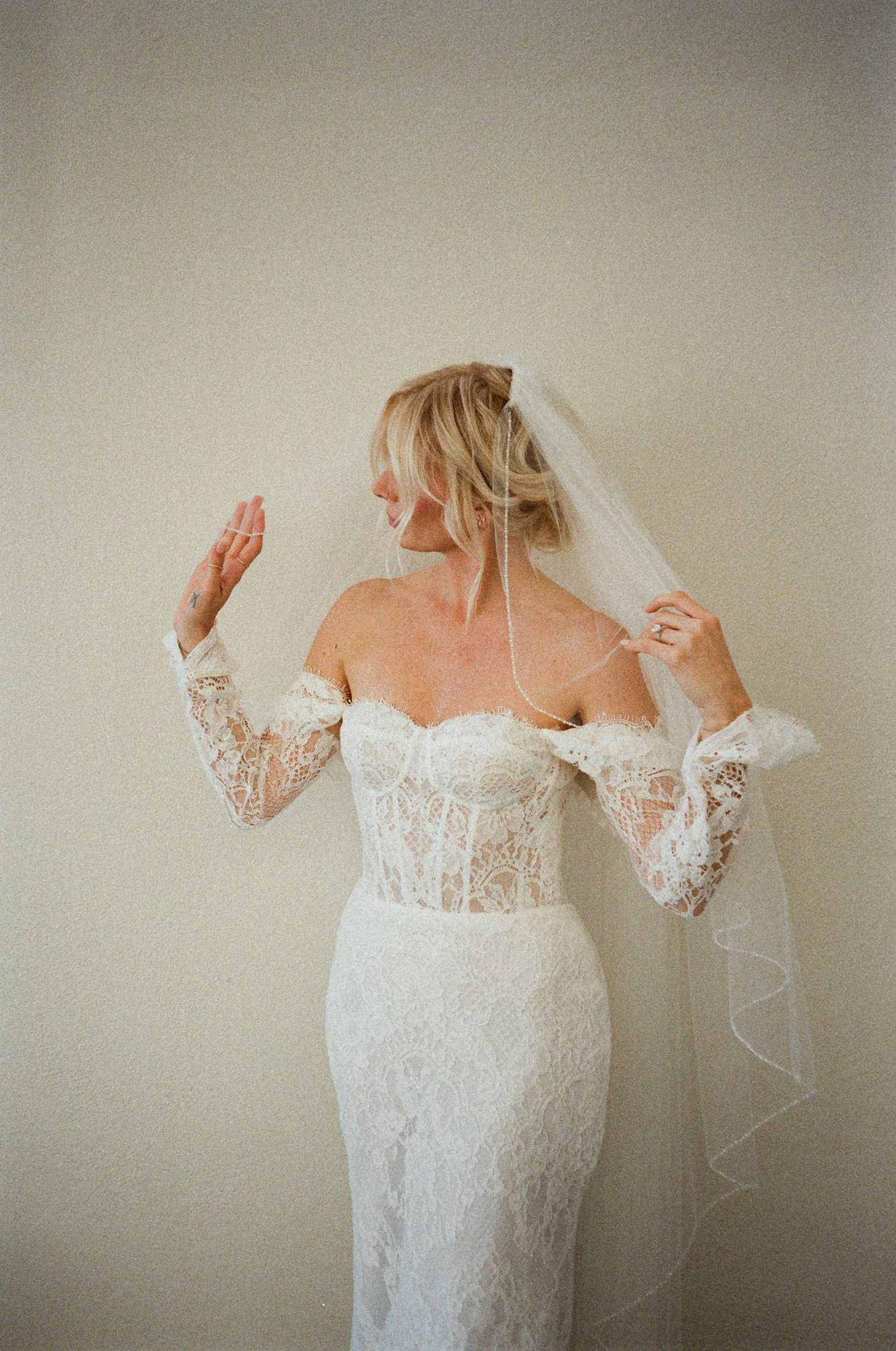 A bride in an off-shoulder lace wedding dress with long sleeves and veil poses against a neutral wall.