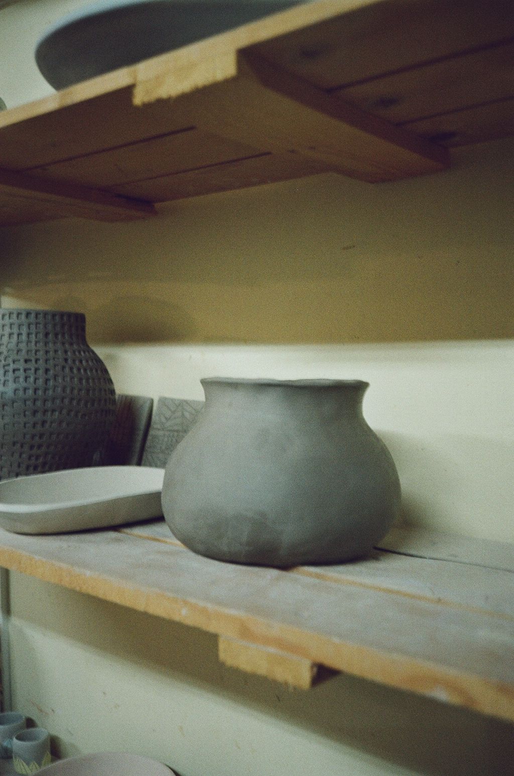 A series of unfinished clay pottery pieces drying on wooden shelves in a dimly lit studio space.