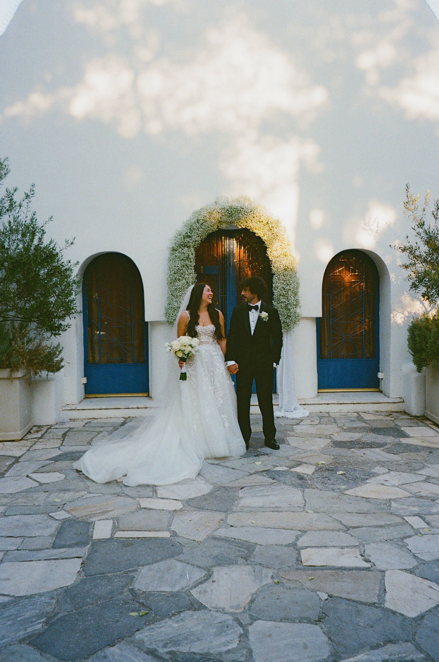 A romantic wedding sequence showing a couple posing against a Mediterranean-style white building with blue windows and stone patio.