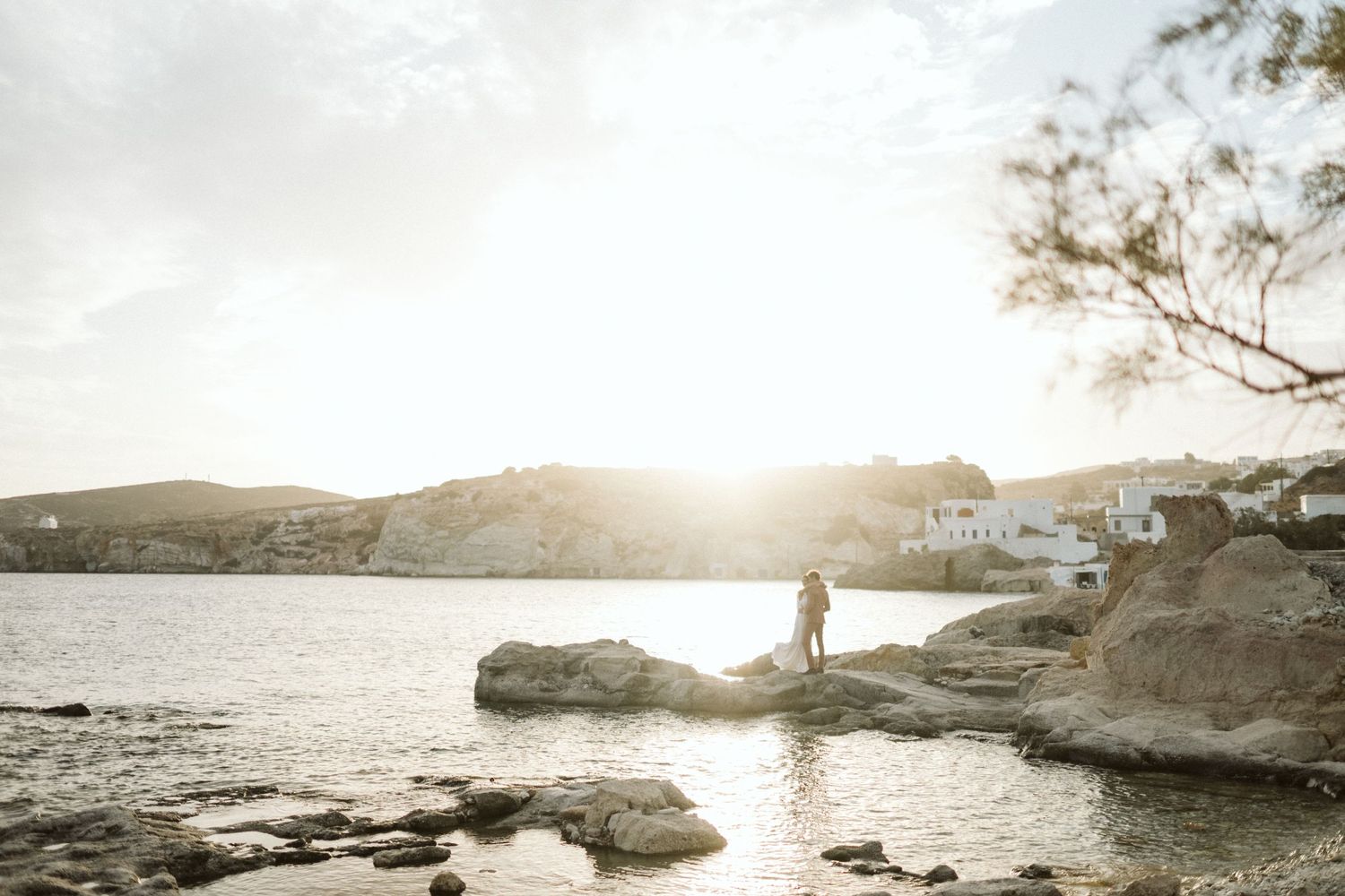 A silhouette stands on rocky coastline during golden hour with sunlight sparkling across tranquil waters.