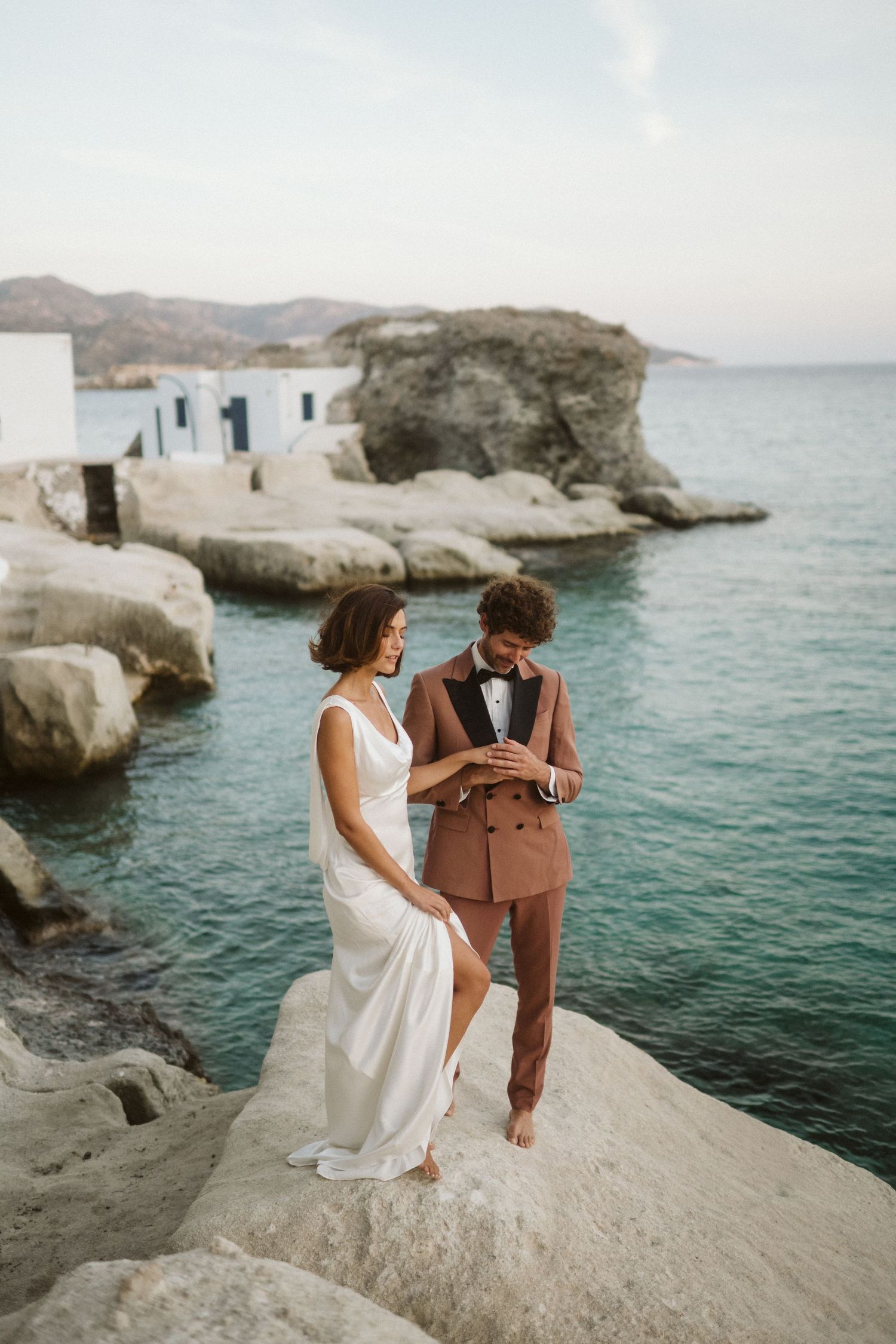 A romantic couple stands barefoot on white seaside rocks overlooking turquoise Mediterranean waters in Greece.