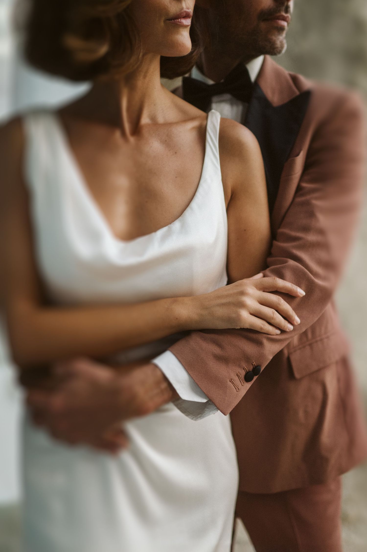 A close-up photo shows a bride in a white silk dress standing close to her partner wearing a brown suit.
