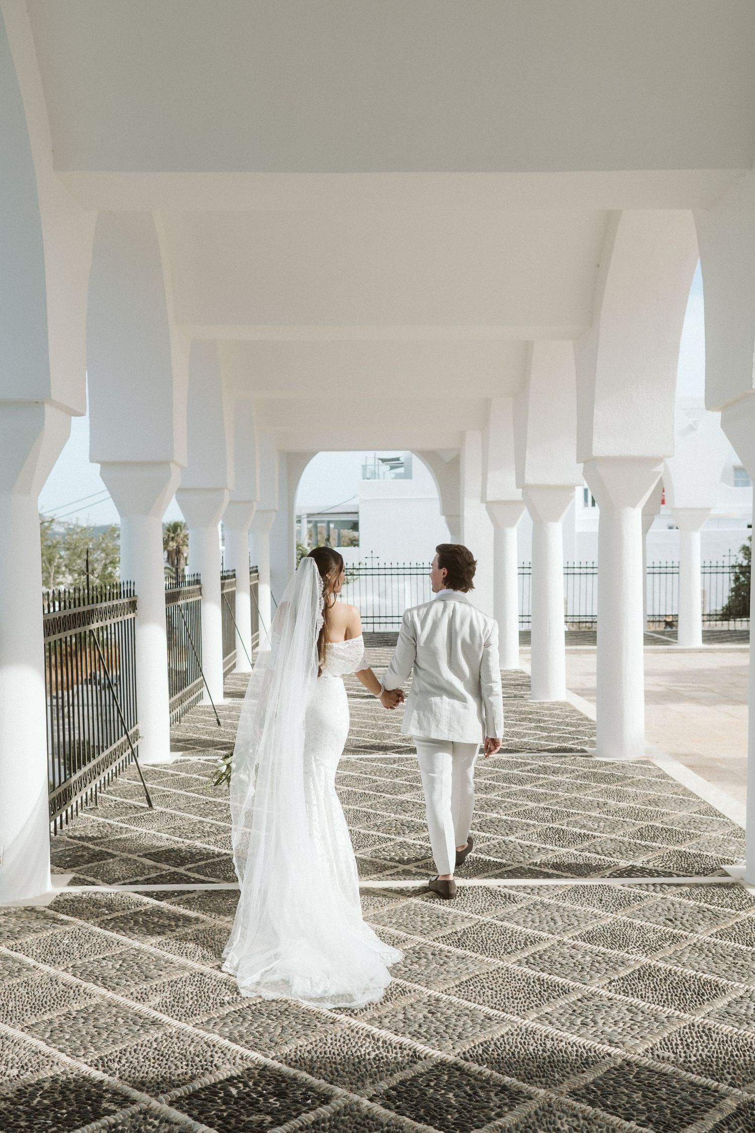 Couple in white wedding attire walks through elegant columned walkway with geometric floor pattern.
