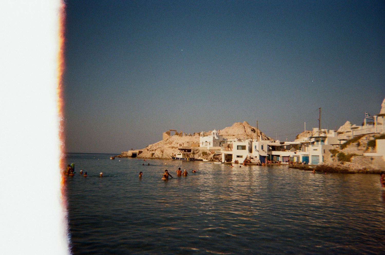 Mediterranean coastal village with white buildings nestled against rocky cliffs beside calm blue waters during sunset.