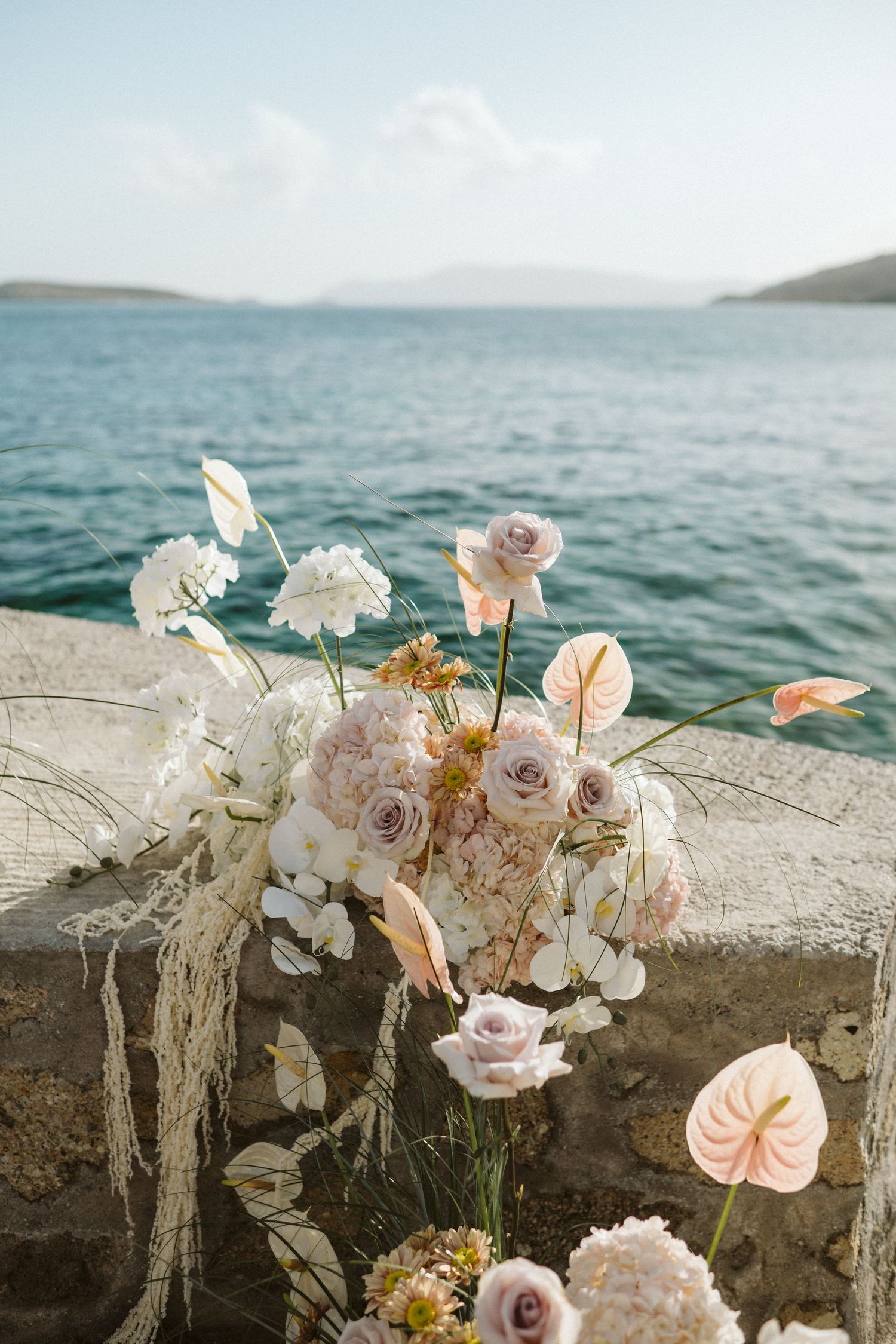 A romantic floral arrangement with pale roses and white flowers against a blue ocean backdrop on a stone wall.