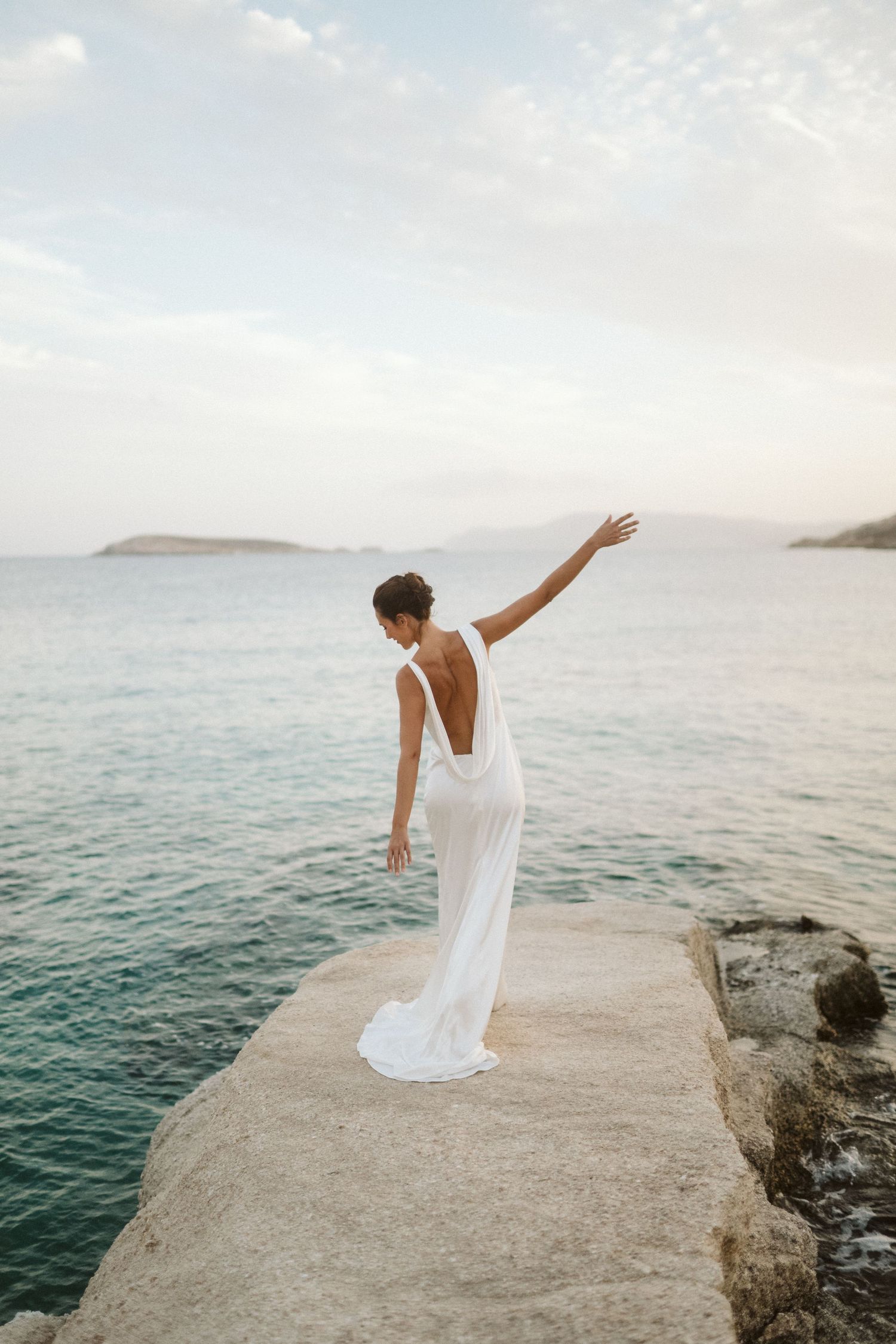 A person in a flowing white dress stands gracefully on a rocky cliff overlooking calm turquoise ocean waters at sunset.