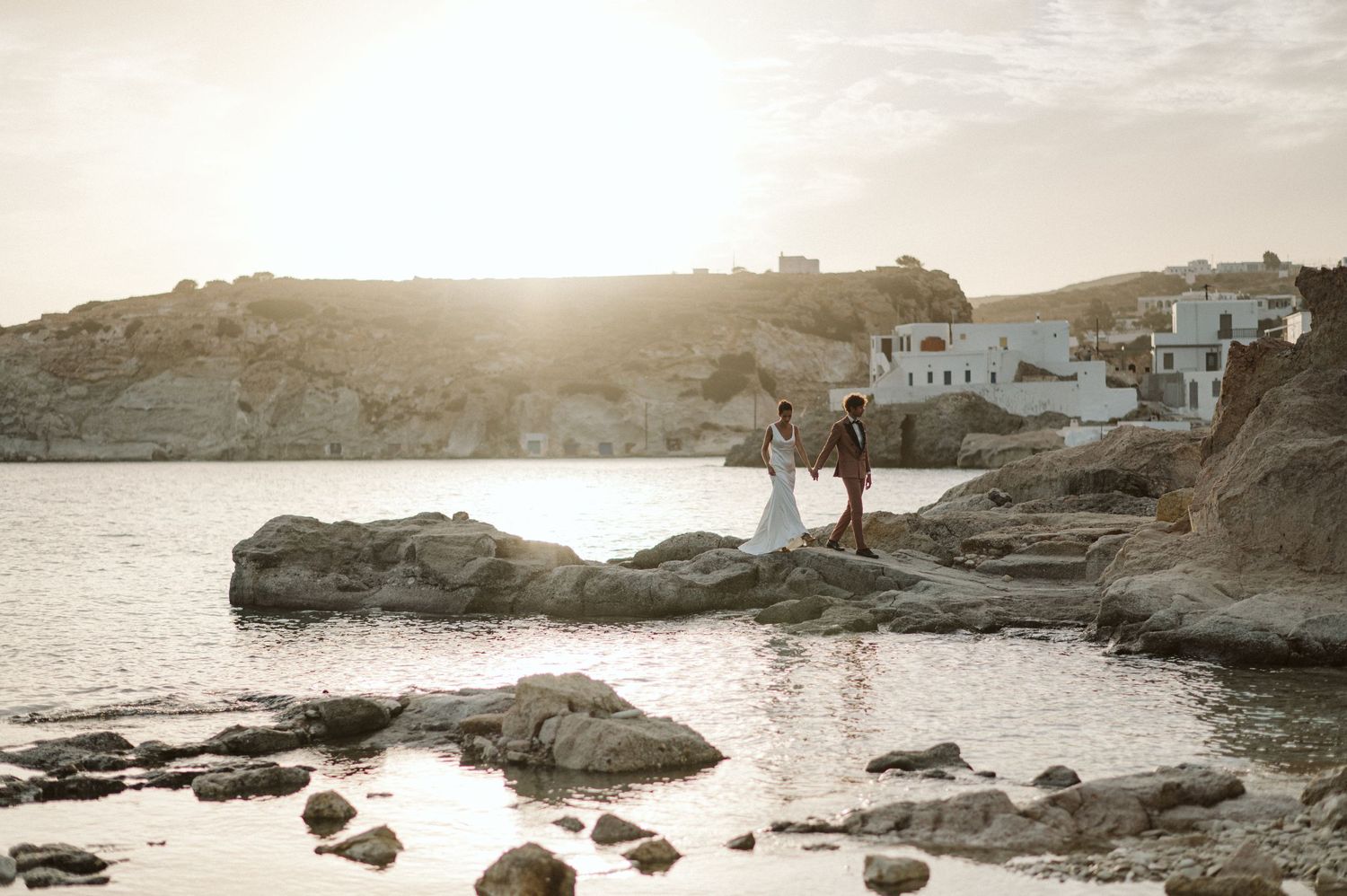 Couple in wedding attire standing on rocky coastal shore at sunset with white Mediterranean buildings in background.