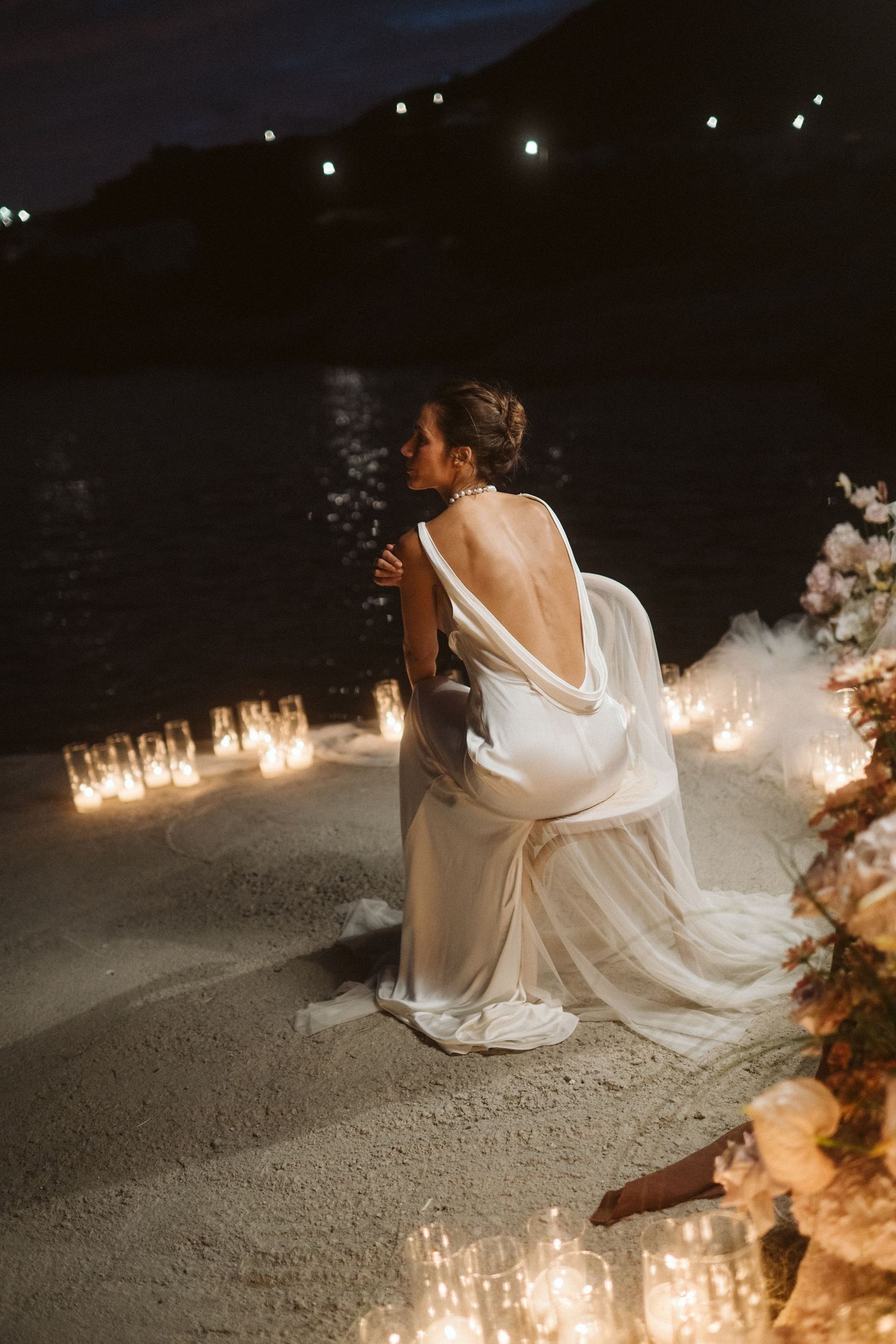 A romantic seaside wedding scene with candlelight illuminating a bride's backless white dress at dusk.
