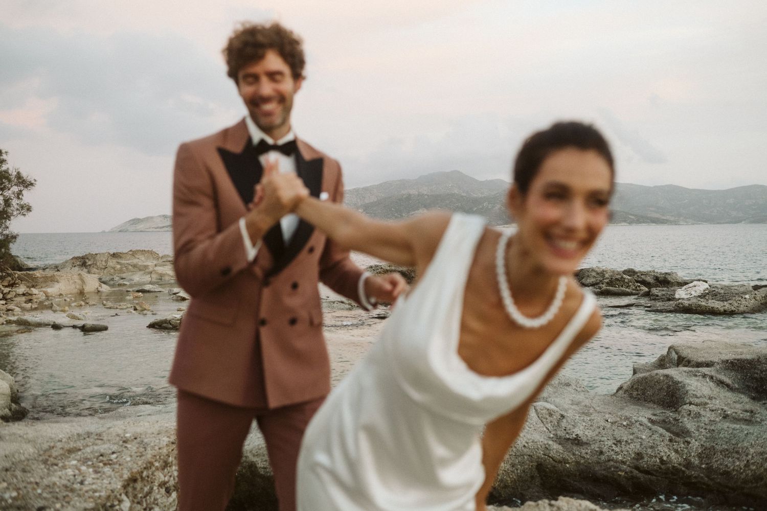 A joyful couple in wedding attire poses on rocky coastal terrain with scenic mountains and water in the background.
