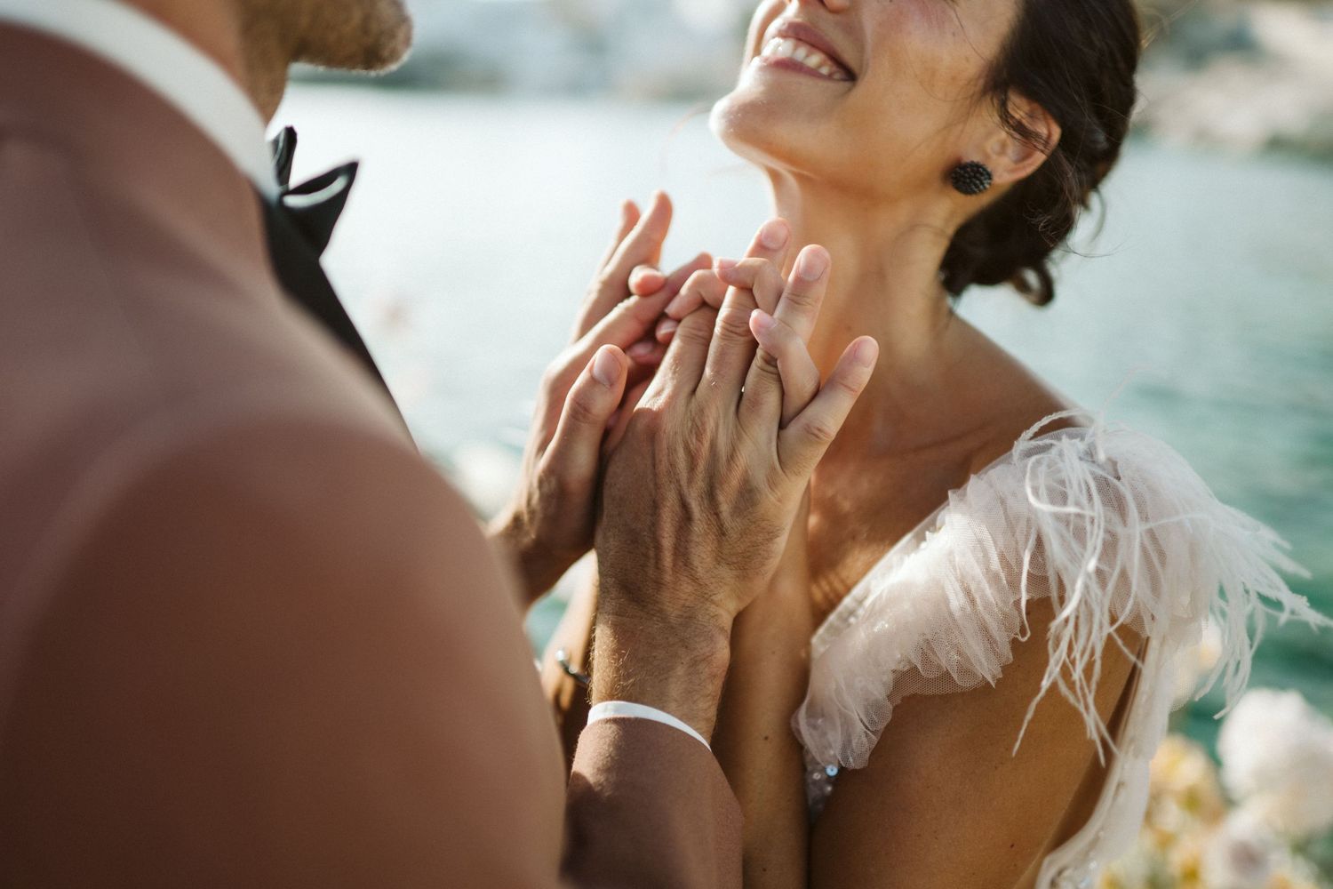 Close up of hands sharing an intimate moment during a waterfront wedding ceremony with white feathered dress detail.