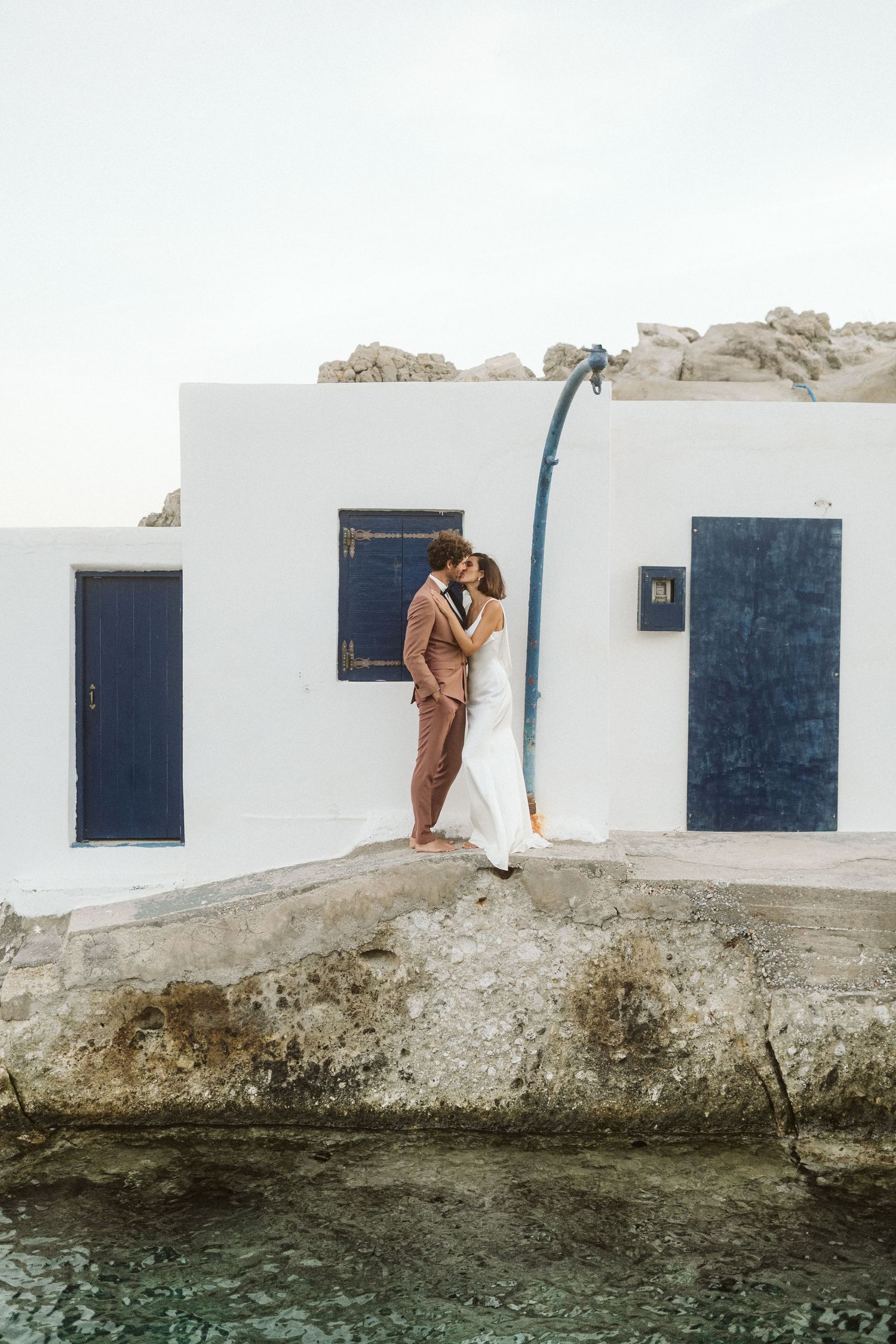 A person in brown clothing stands against a white Mediterranean building with blue doors next to turquoise waters.