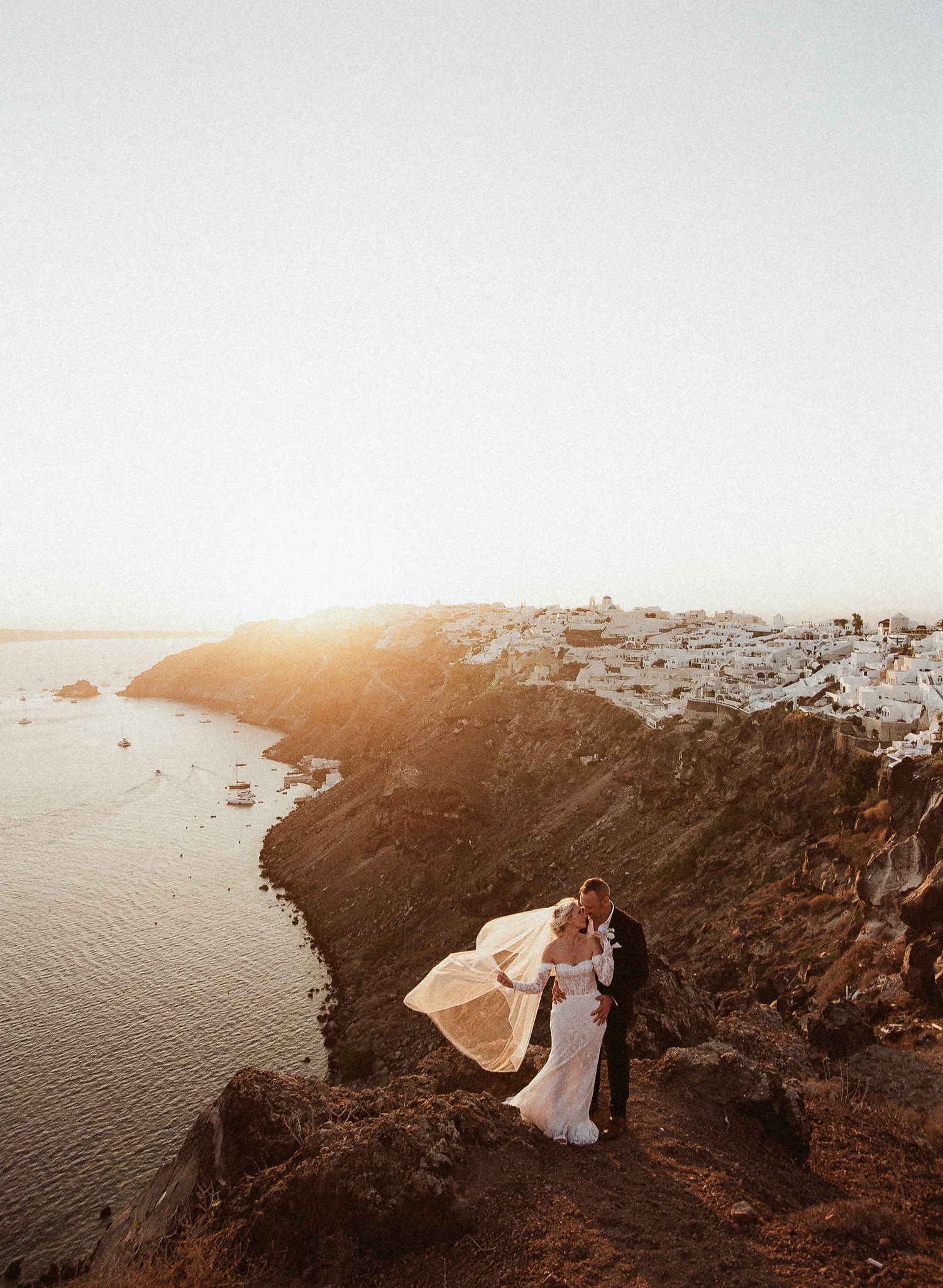 A romantic sunset silhouette of a couple standing on a cliffside overlooking the Mediterranean Sea in Santorini, Greece.