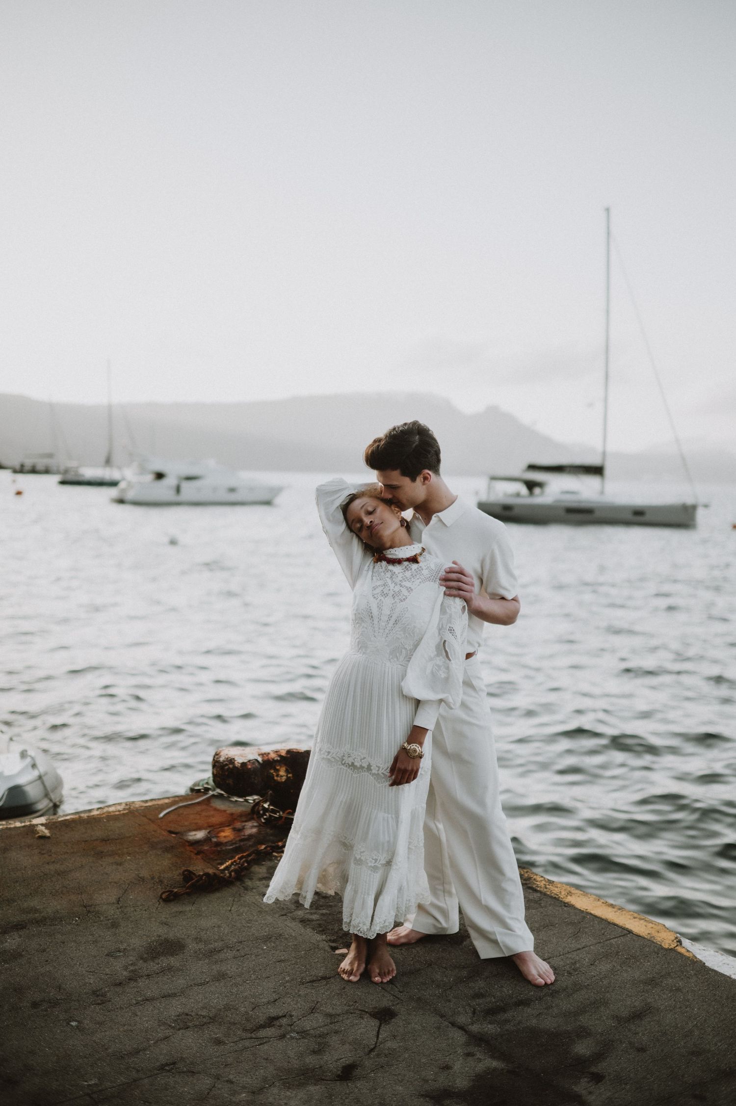 Romantic coastal wedding photo with couple embracing by the water with sailboats in the background.