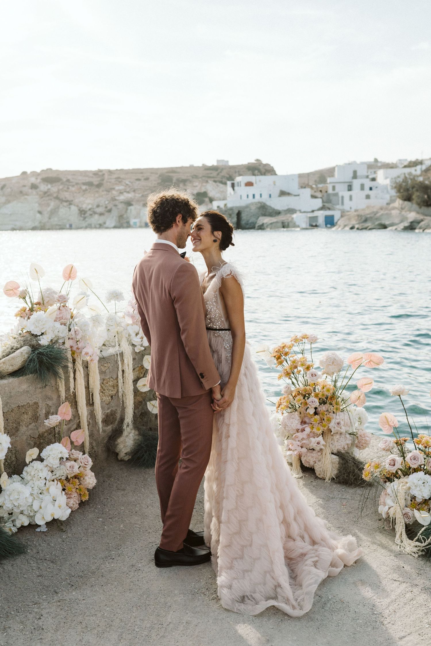 A couple shares a romantic moment by the sea, surrounded by floral arrangements and white Mediterranean buildings.