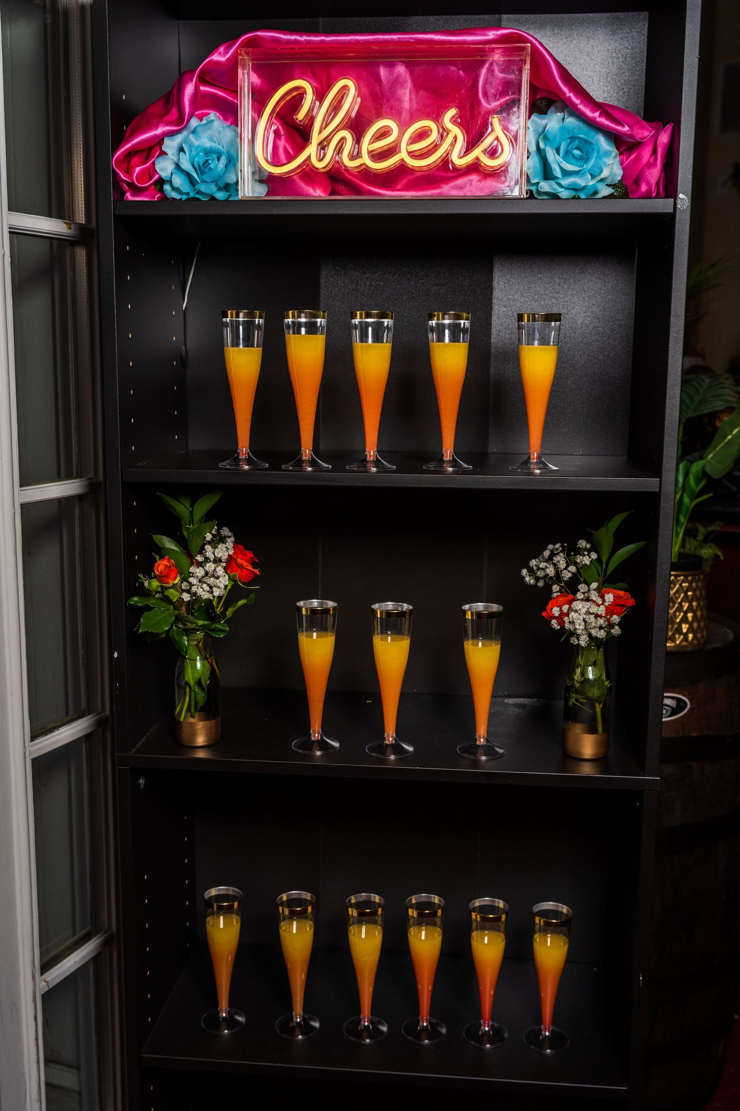 Black shelving unit displays rows of orange mimosas with a neon Cheers sign on top.