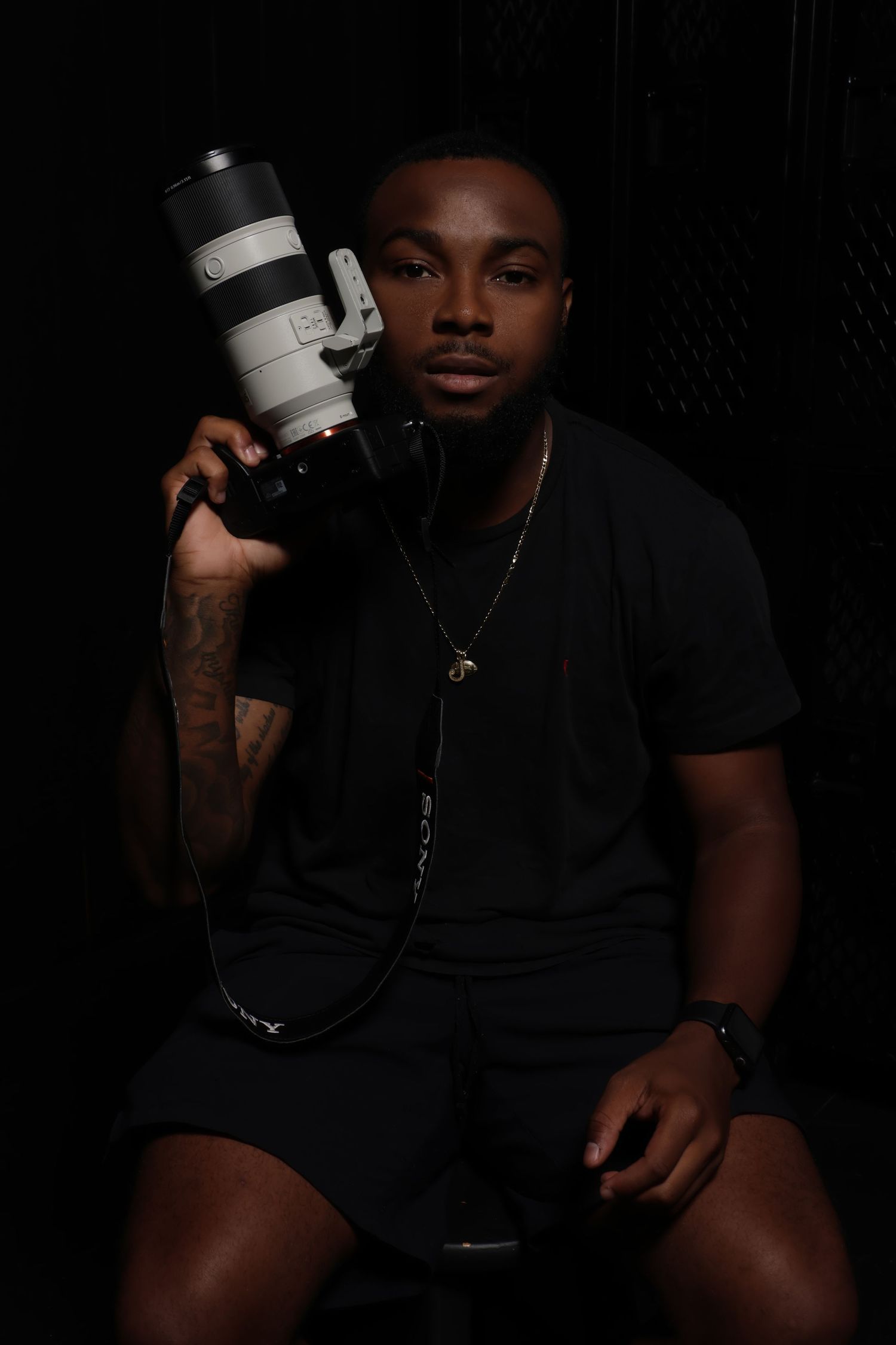 Young Black male photographer posing with Sony telephoto lens camera in dark studio setting.