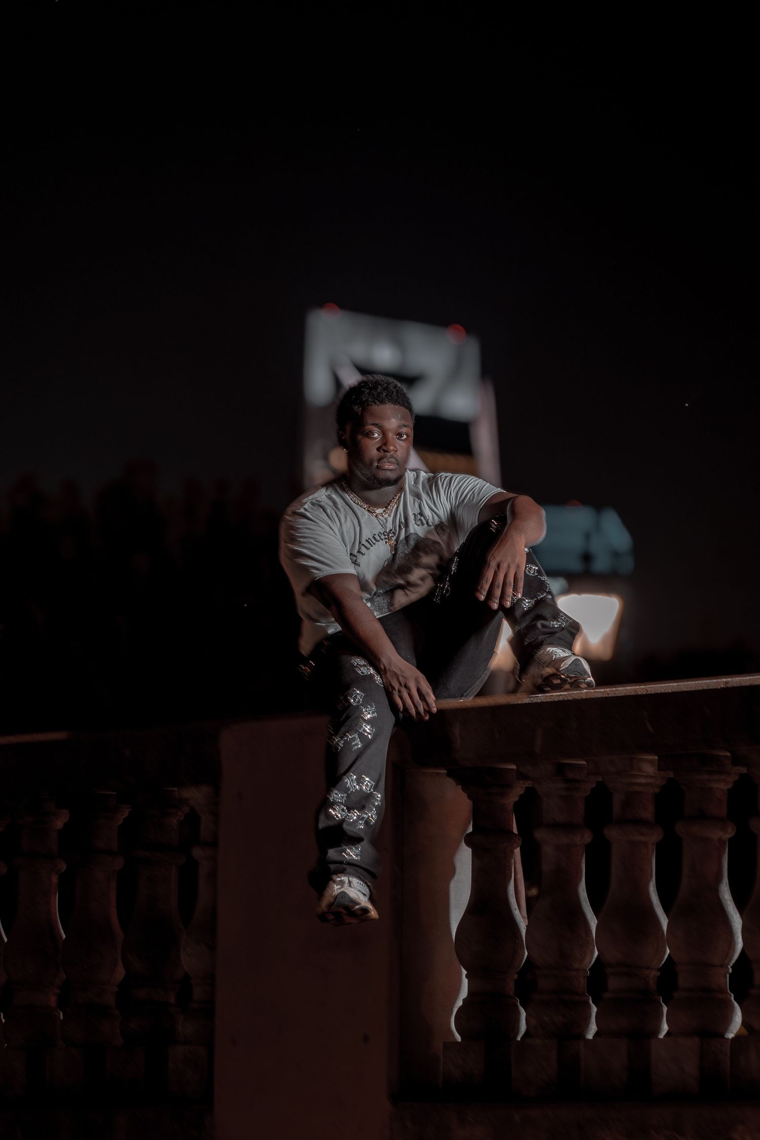 Young man sitting on a stone balustrade at night wearing camo pants and white tee.