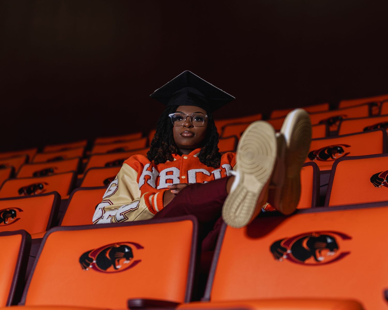 Graduate in cap and HBCU jacket sitting among orange stadium seats with feet up.