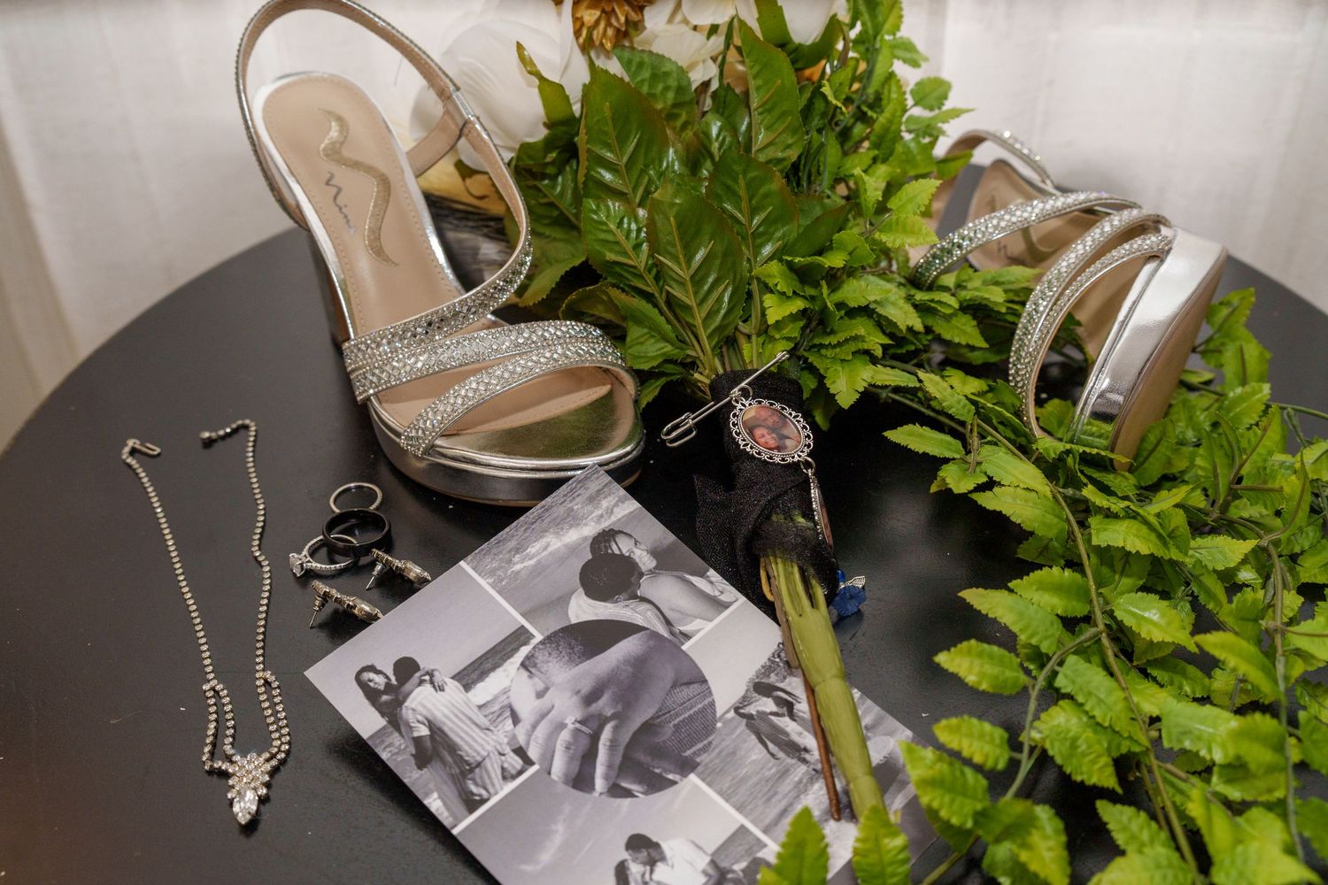 Bridal accessories flat lay featuring silver heels, jewelry, watch, and greenery on a dark table.