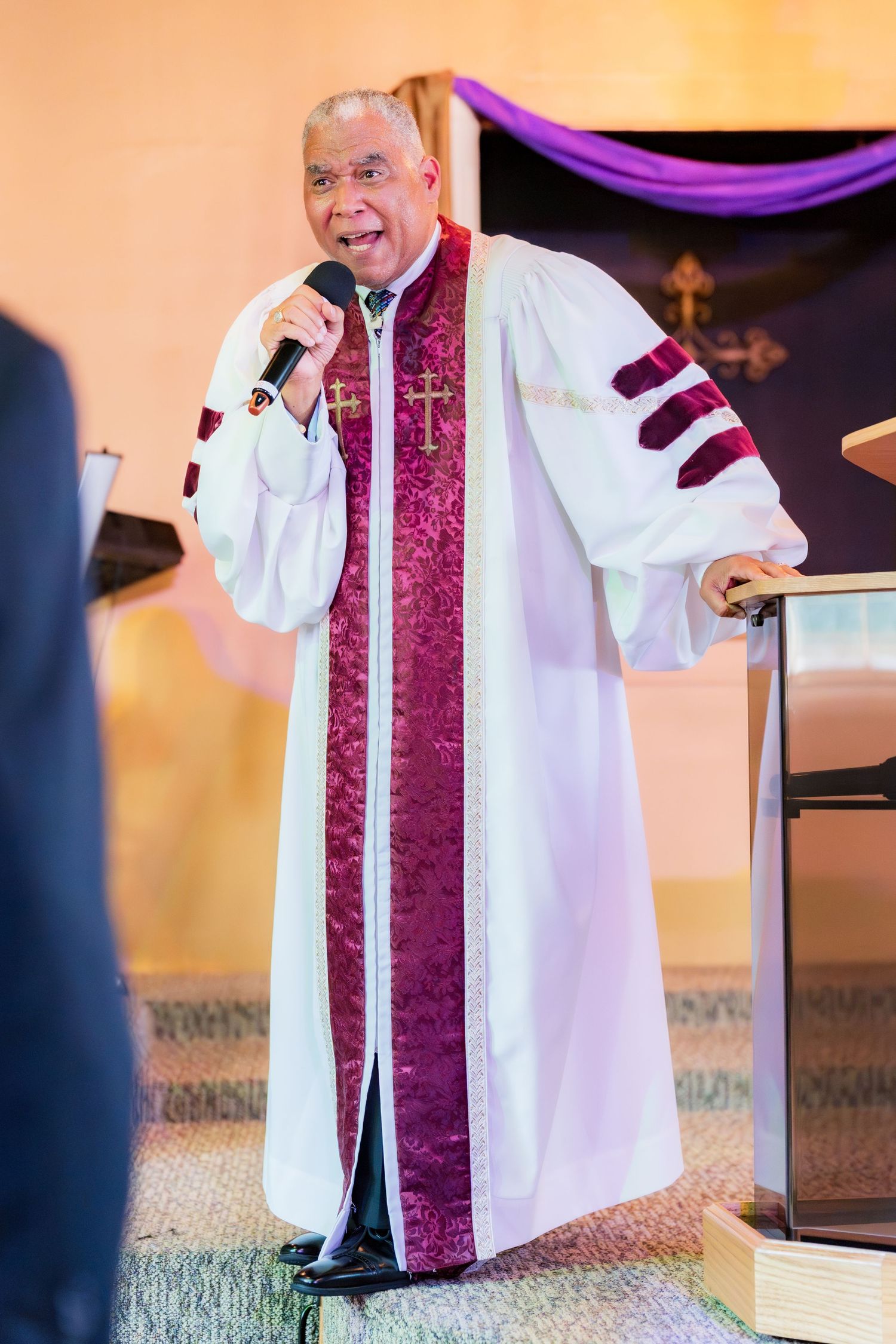 Pastor in white and burgundy ceremonial robes speaking at podium during church worship service.