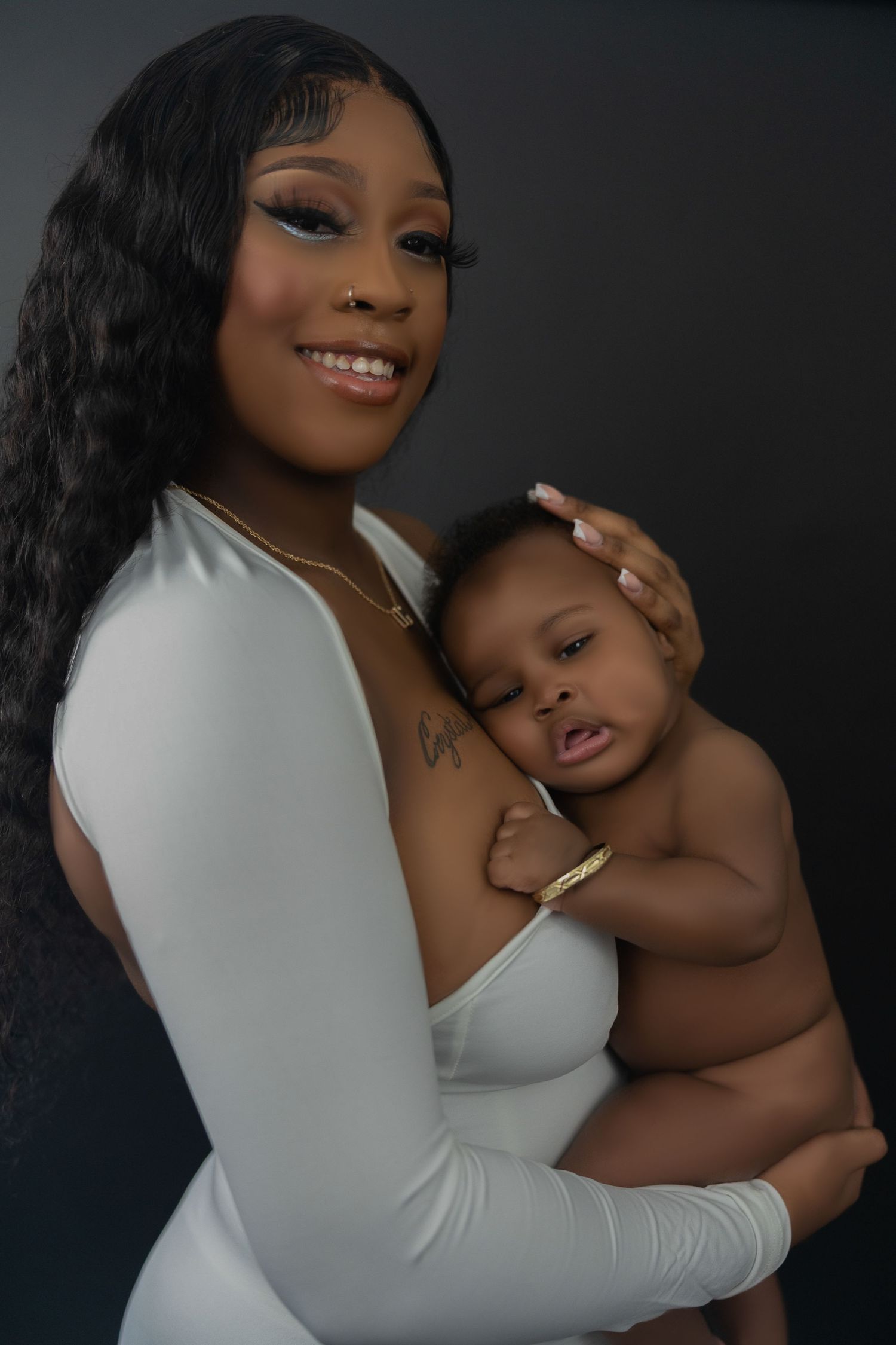 Young mother in white bodysuit cradles sleeping newborn baby in dark studio portrait.