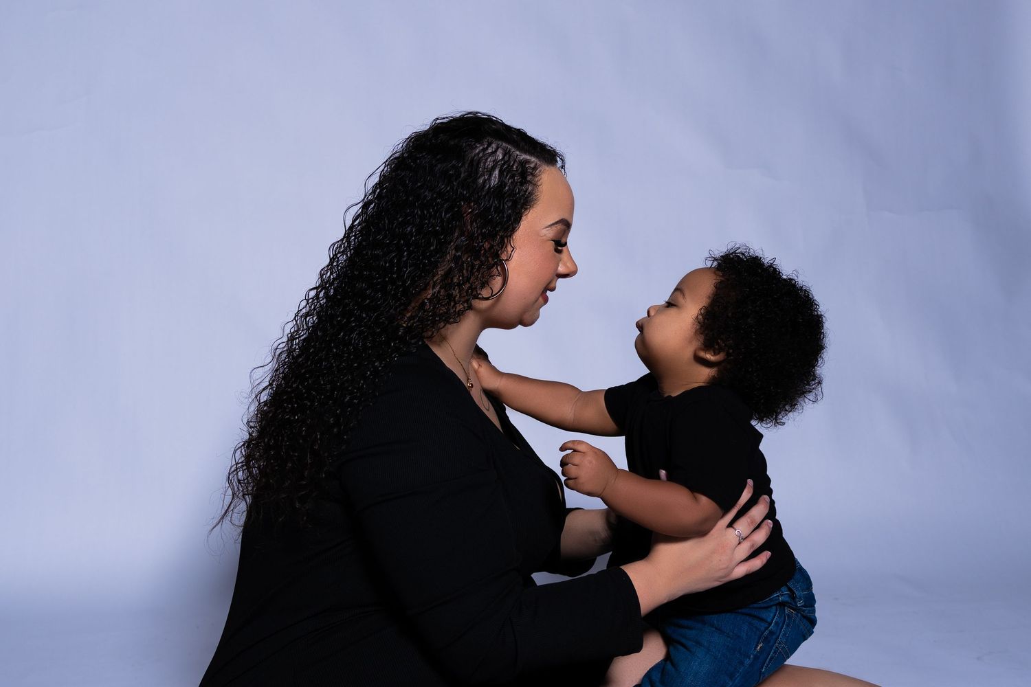 Mother with curly hair gazes lovingly at toddler in black outfits against gray background.