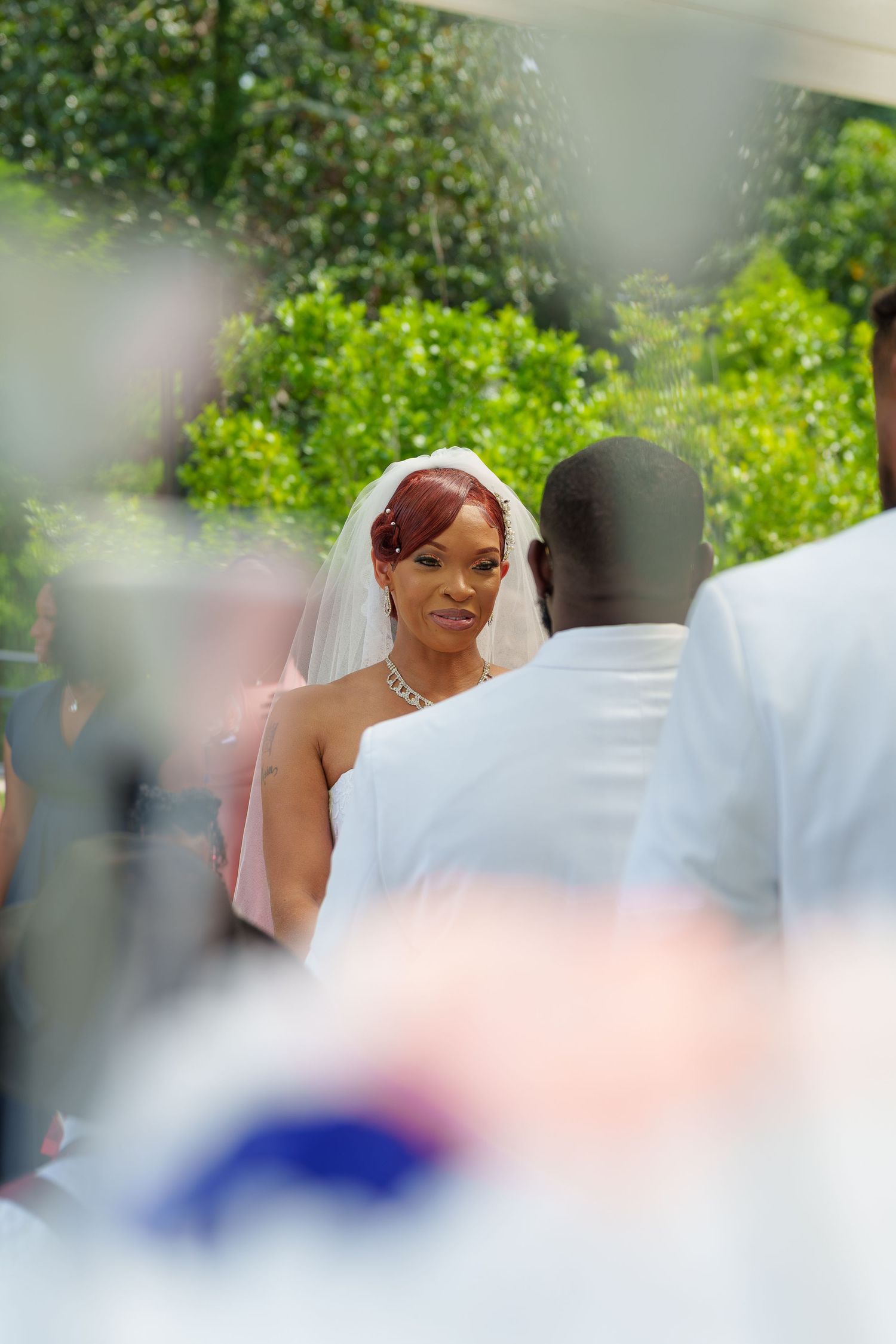 Bride in white veil gazing at groom during outdoor wedding ceremony surrounded by greenery.