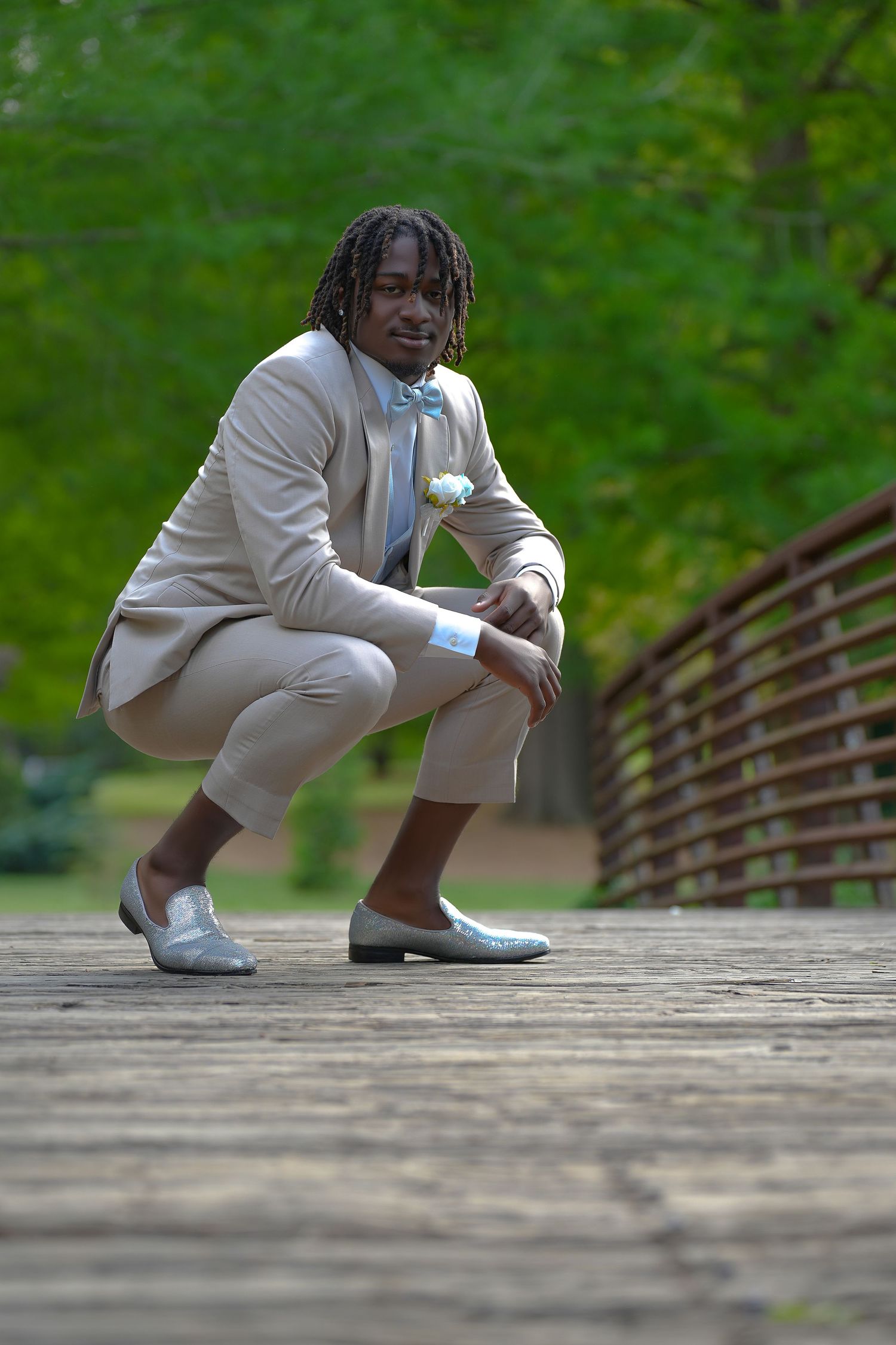 Young man in tan prom suit with boutonniere crouching on wooden bridge in green park.