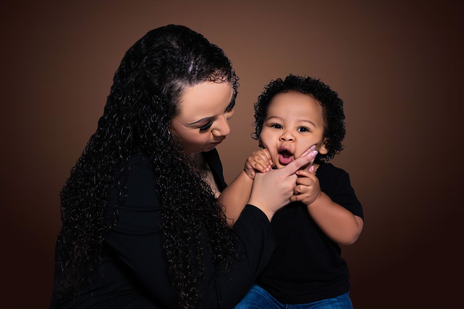 Woman with curly hair smiling at laughing baby during studio portrait session.